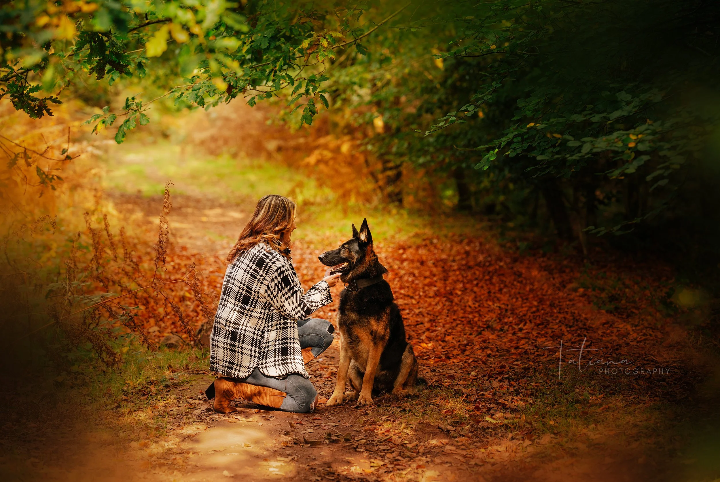 A woman kneeling on a forest trail holding a German Shepherd dog by the collar amid autumn foliage.