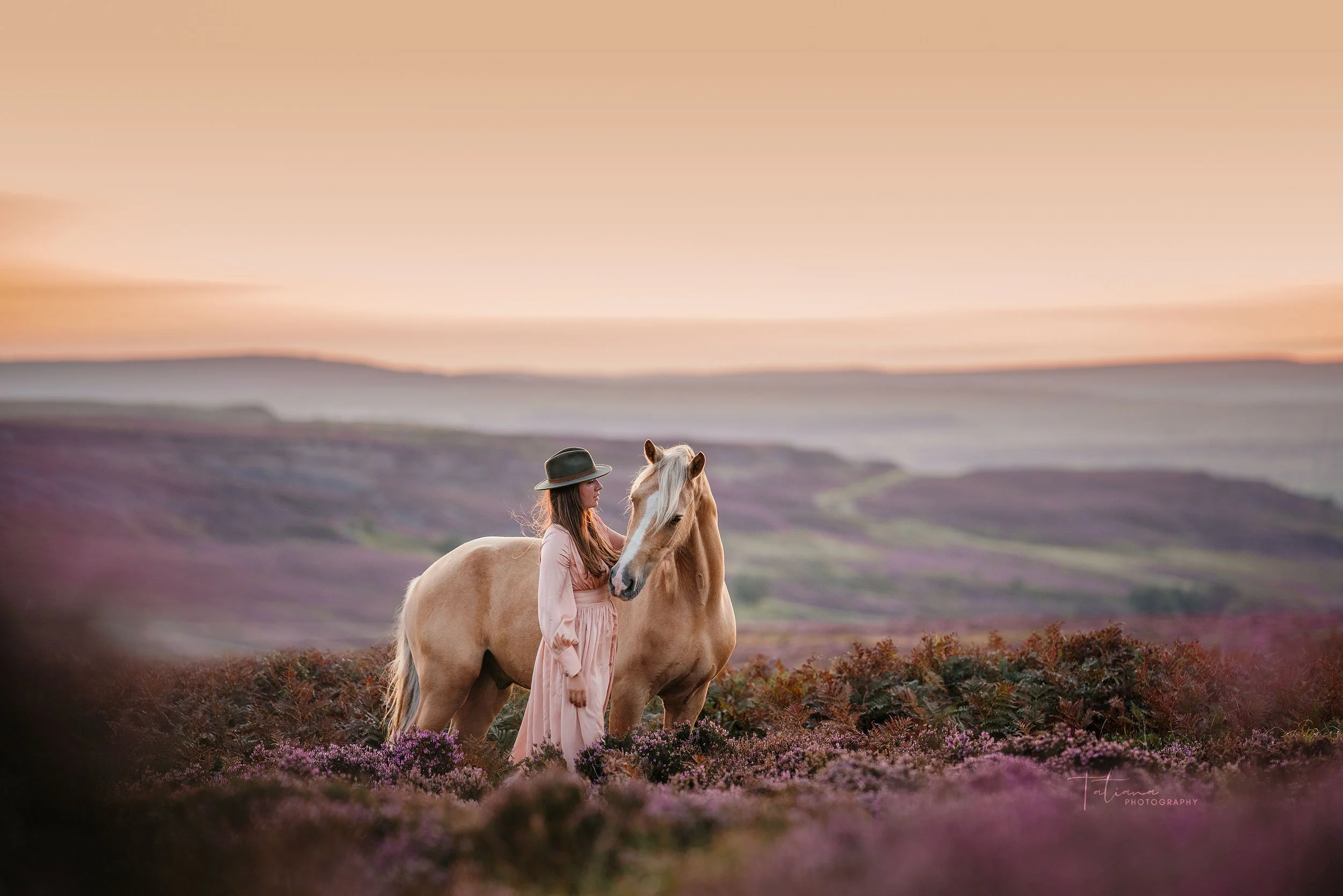 A woman in a pink dress and black hat standing next to a light-colored horse in a landscape with rolling hills and purple flowering plants at sunset or sunrise.