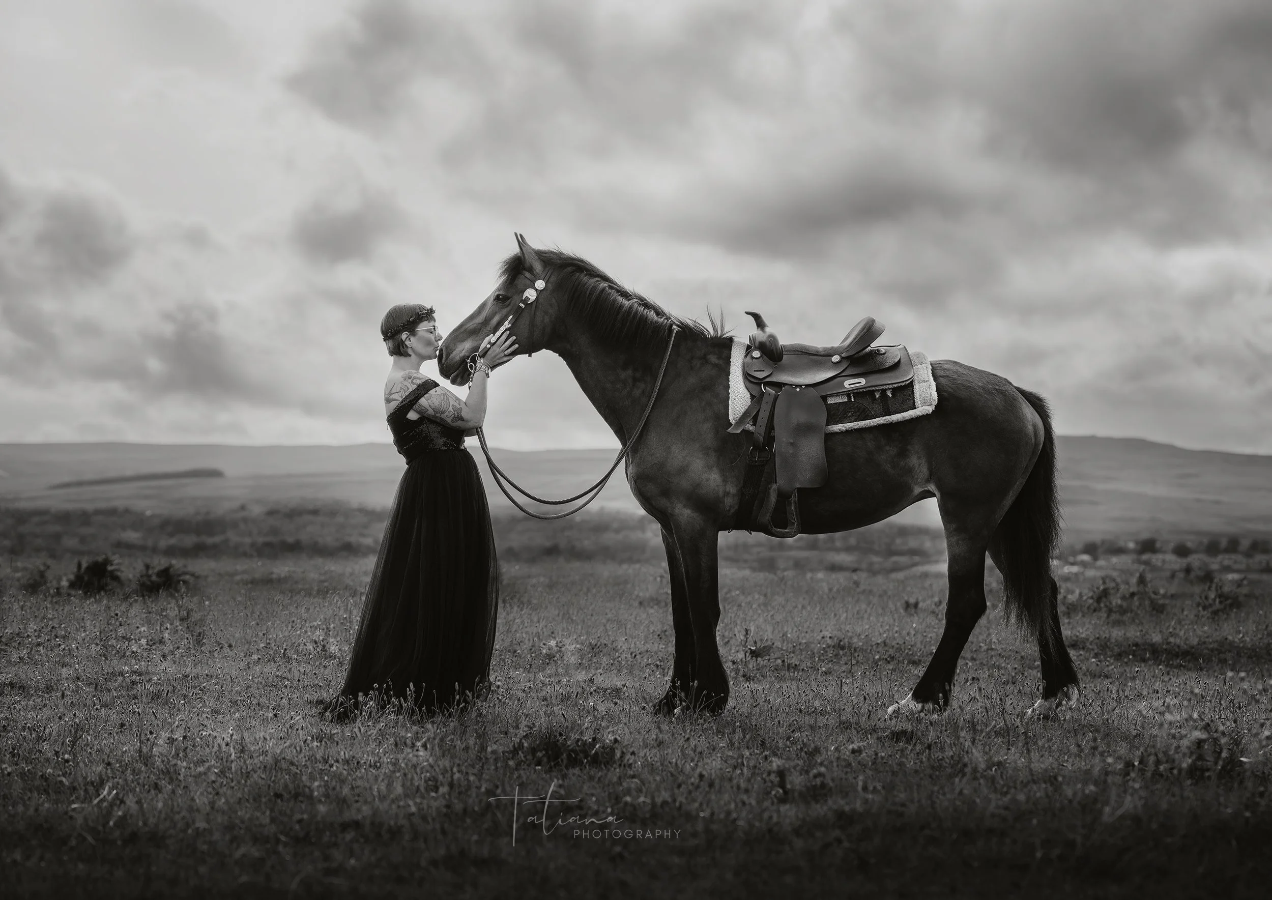 A woman in a long black dress and glasses touching a horse's face in a field under cloudy skies.