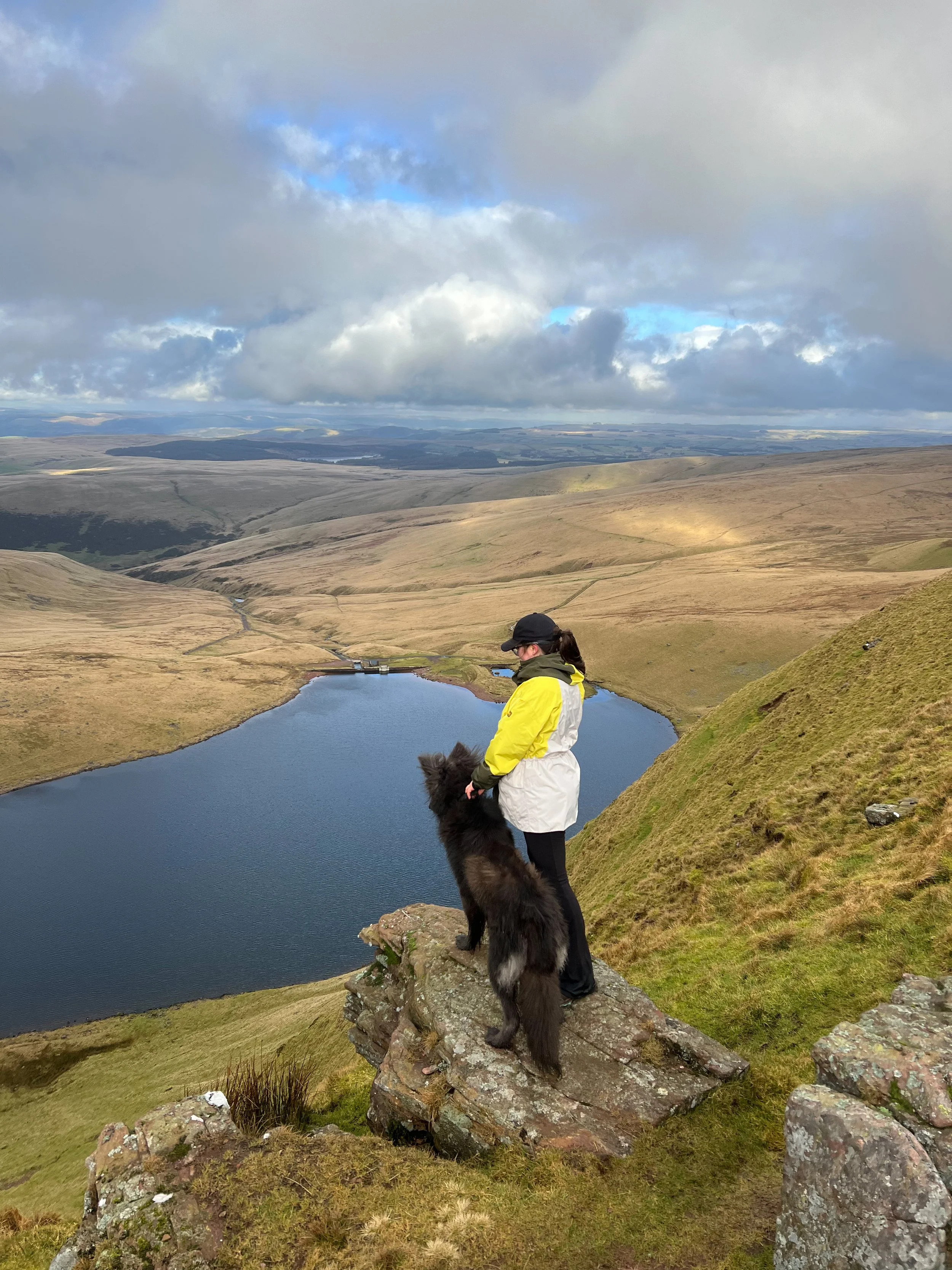 A woman with a black dog standing on a rock ledge overlooking a large lake in a hilly landscape under a cloudy sky.