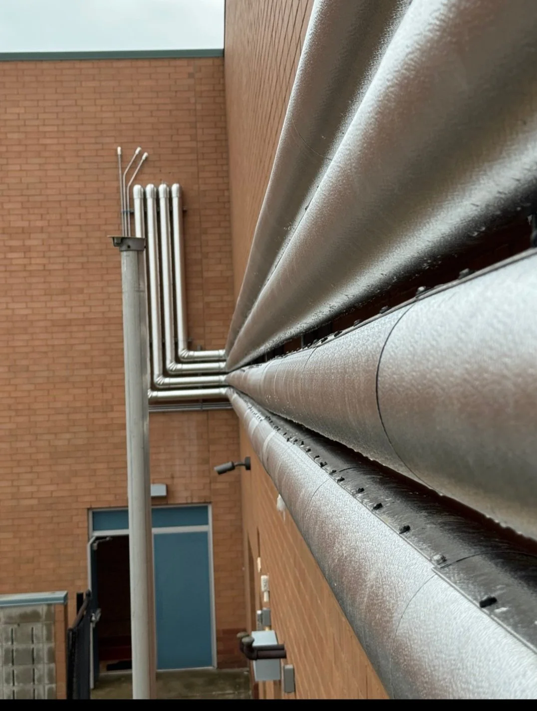 Close-up view of metallic pipes running along the side of a brick building, with a door and window visible in the background.