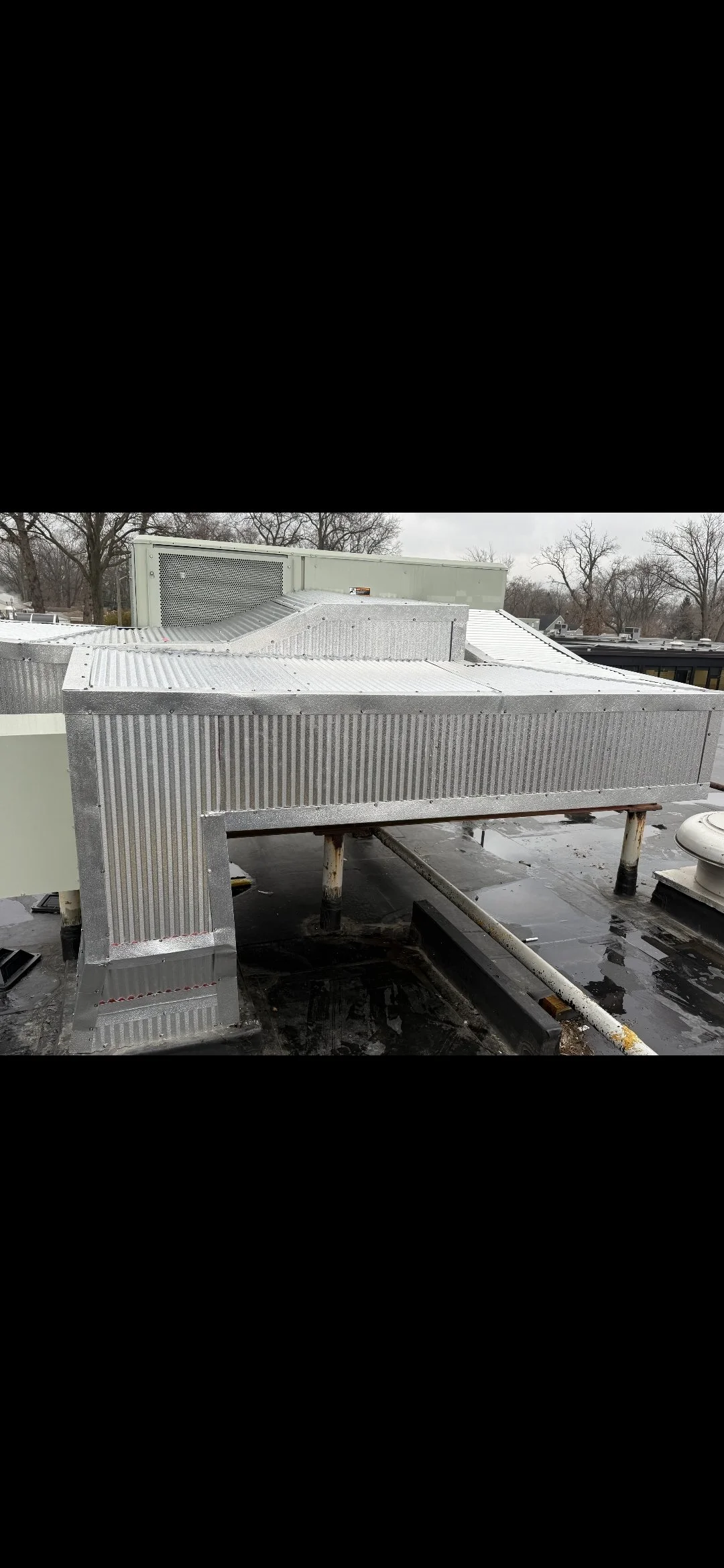 Metal rooftop HVAC units and ductwork with trees and overcast sky in the background.