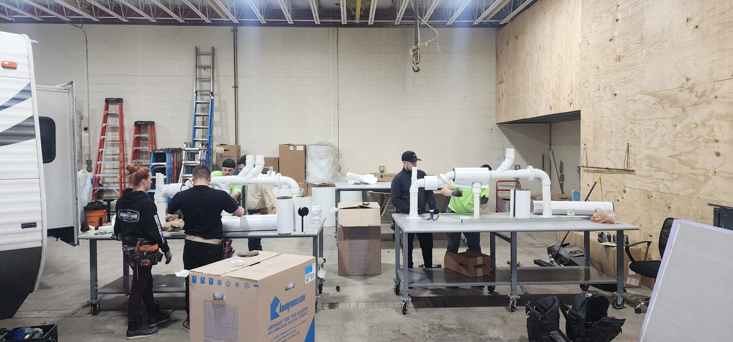 People assembling large white PVC pipes in a warehouse with ladders and cardboard boxes around.