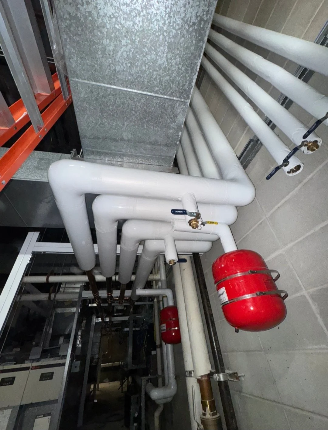 Network of white pipes and valves with red fire suppression system cylinders in an industrial or commercial facility.