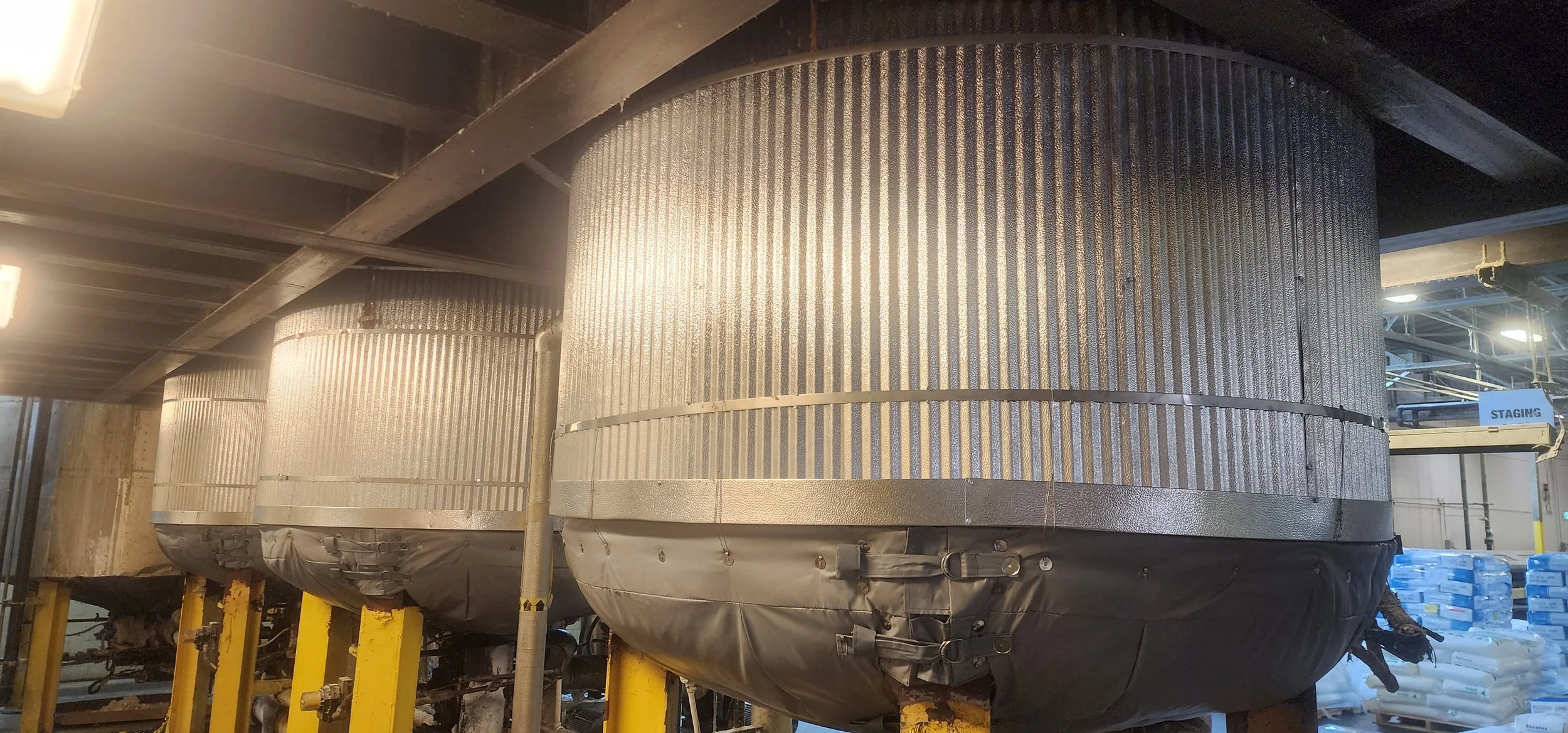 Industrial cooling towers with metallic surfaces and black insulation at the bottom, located in an industrial setting with pipes and storage piles nearby.