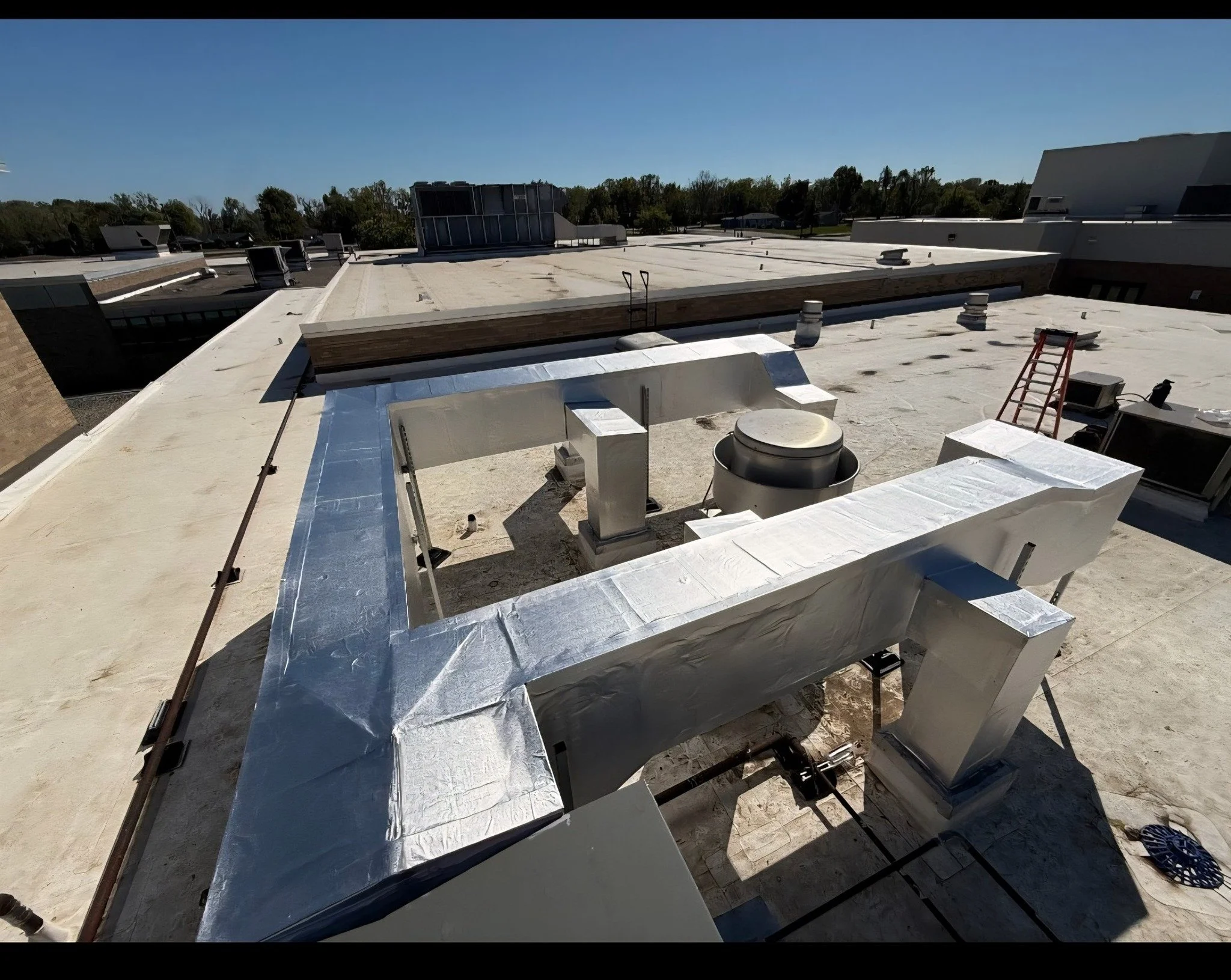 Rooftop with HVAC ventilation ductwork wrapped in silver insulation, ladder, and vents under a clear blue sky.