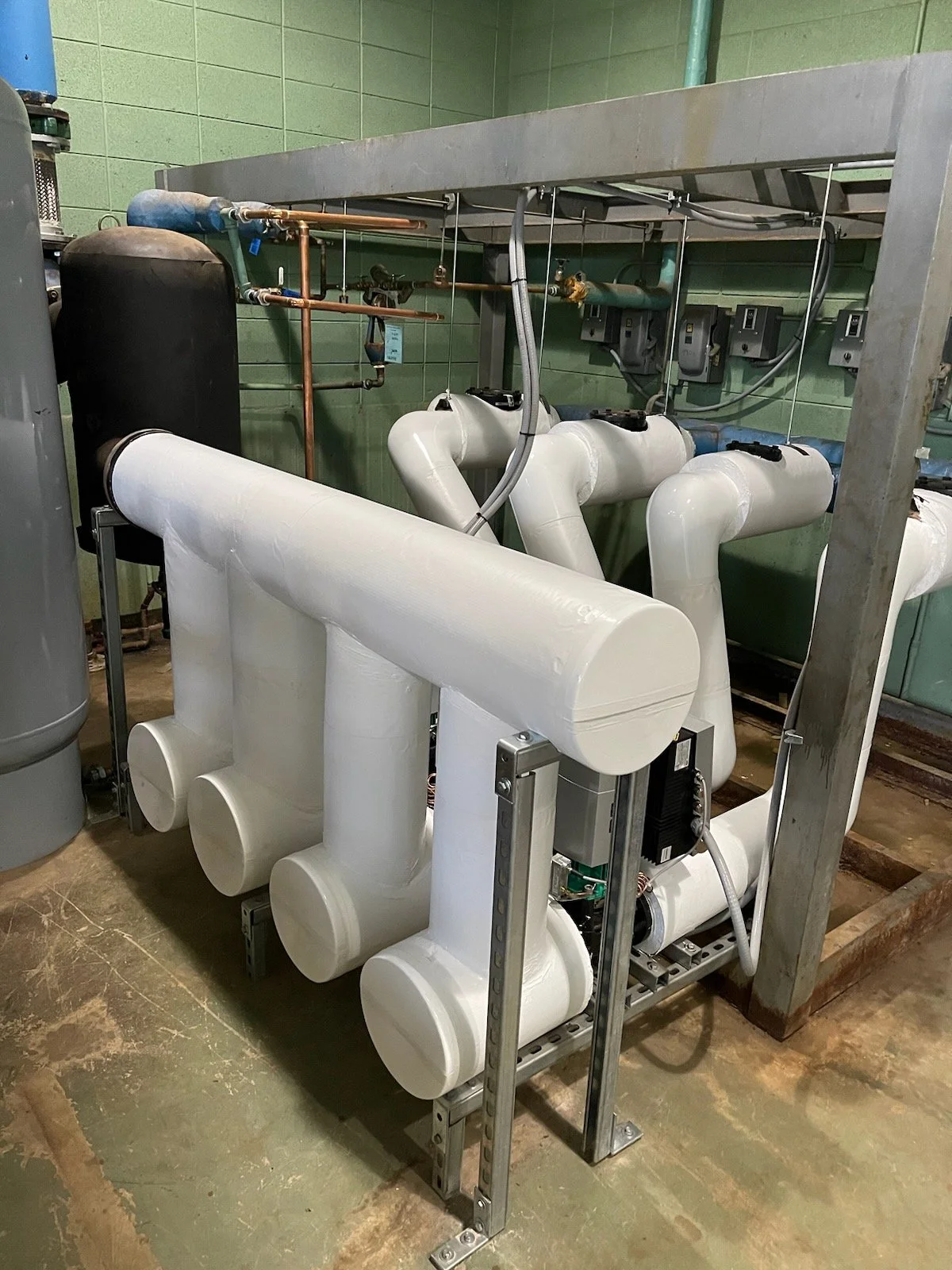 Industrial equipment with large white insulated pipes connected to various valves and control boxes, situated in a room with green tiled walls.