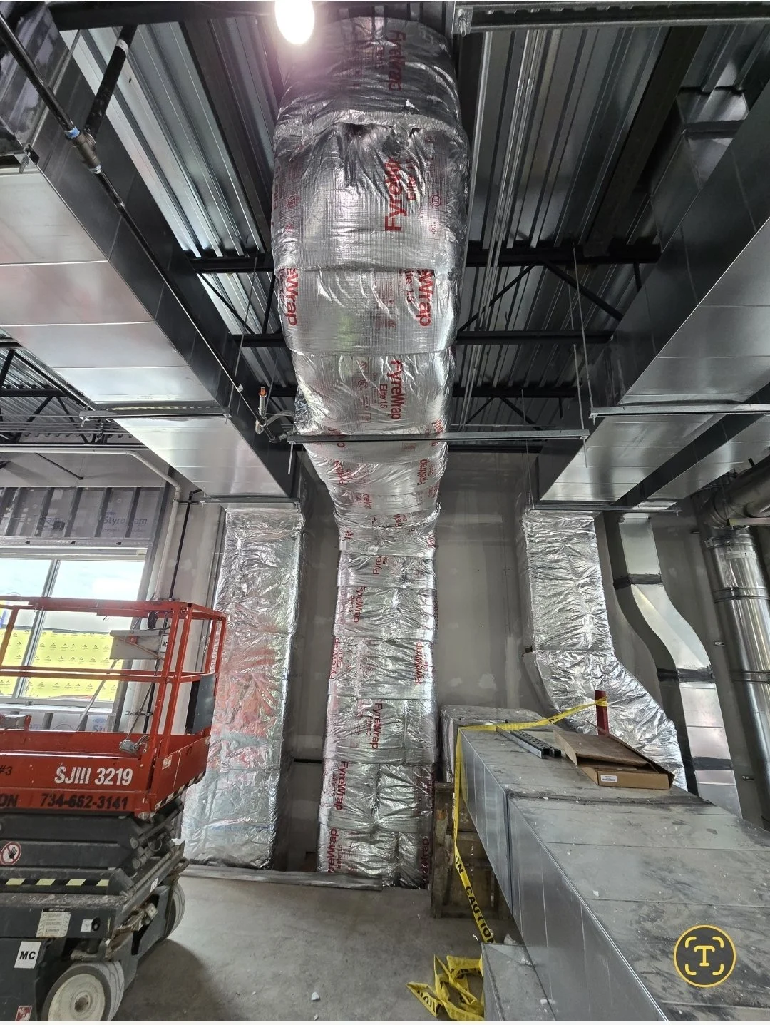 Industrial interior with uncovered ductwork and ventilation pipes, a red scissor lift, construction tools, and caution tape.