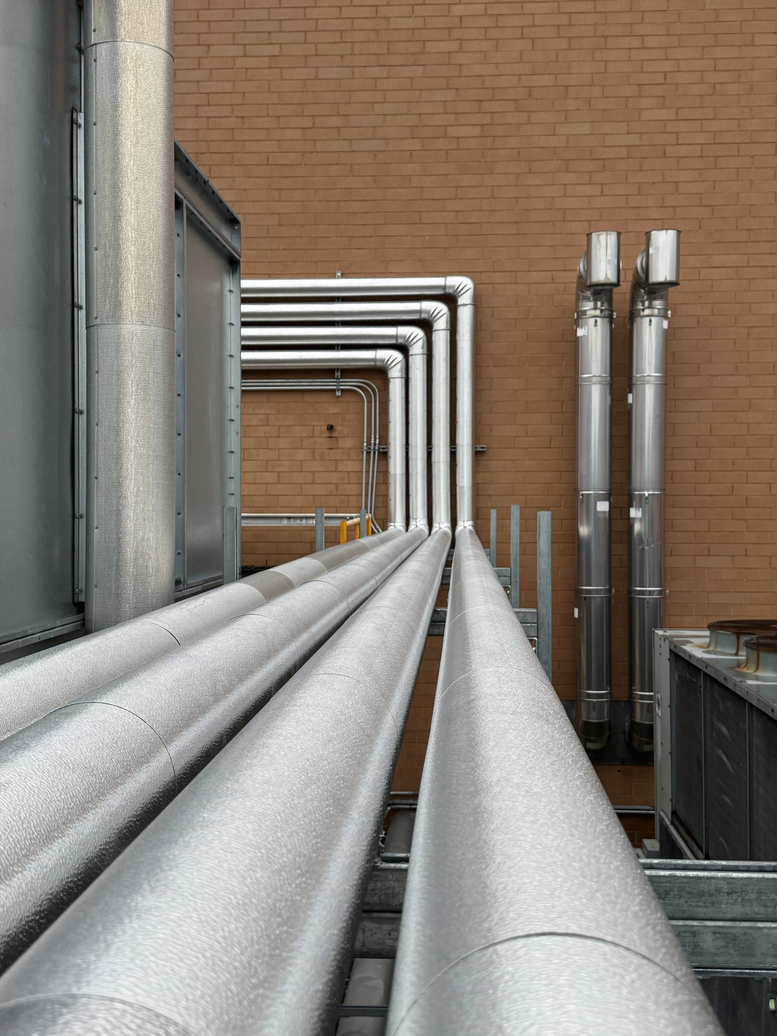 Multiple large metallic pipes running parallel along a building wall with a brick exterior.