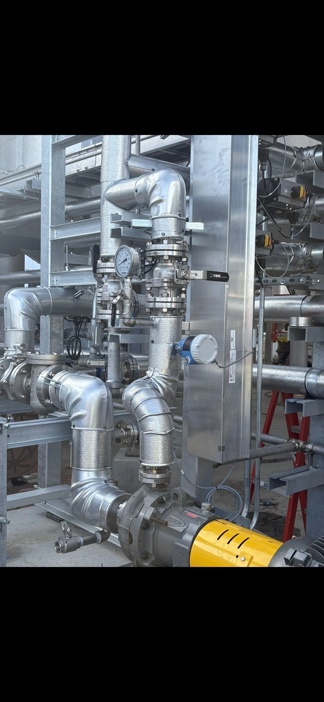 Industrial piping system with metallic pipes, valves, gauges, and a motor, installed in a factory or plant setting.