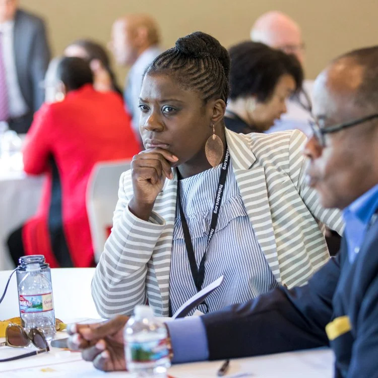 Shaya Gregory Poku with braided hair and a striped blazer sits at a table during a conference, appearing thoughtful with her hand on her chin. Other conference attendees are visible in the background.