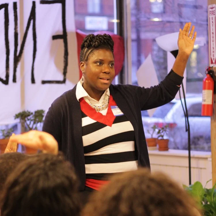 Shaya Gregory Poku standing in front of a group, speaking and raising her hand in a room with large windows, plants, and a fire extinguisher.