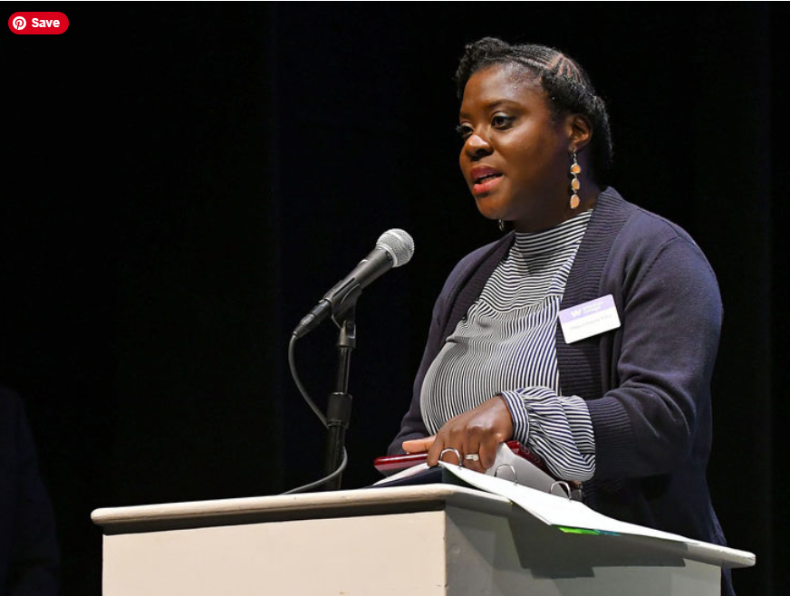 Shaya Gregory Poku speaking at a podium with a microphone, wearing a dark blazer, striped shirt, and earrings, holding a notepad and pen.