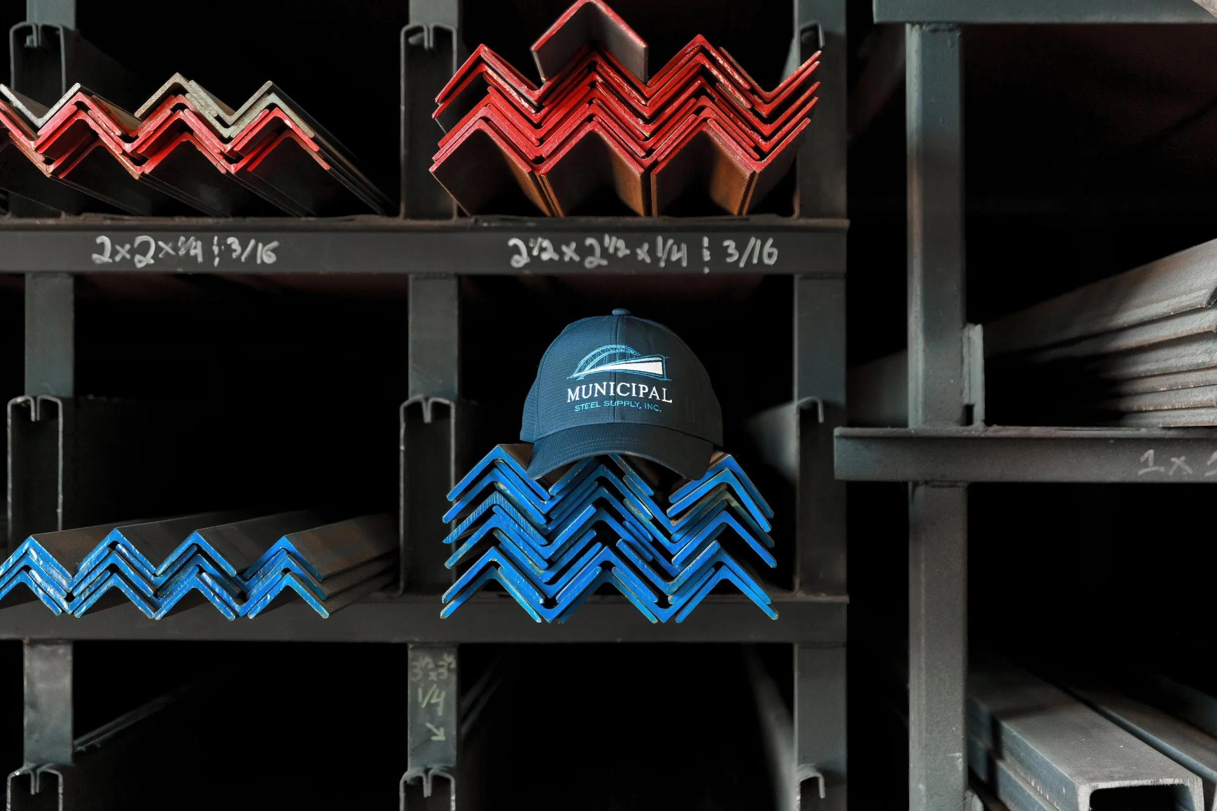 Steel shelves holding various metal angle bars, some painted red and blue, with white handwritten dimensions written on the shelves, and a blue cap with a logo below the bars.