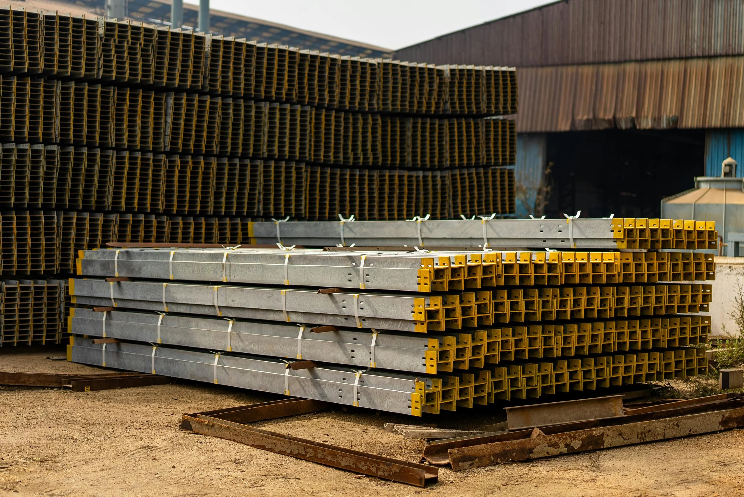 Metal and wood construction materials stacked outdoors in front of a building with a rust-colored wall.