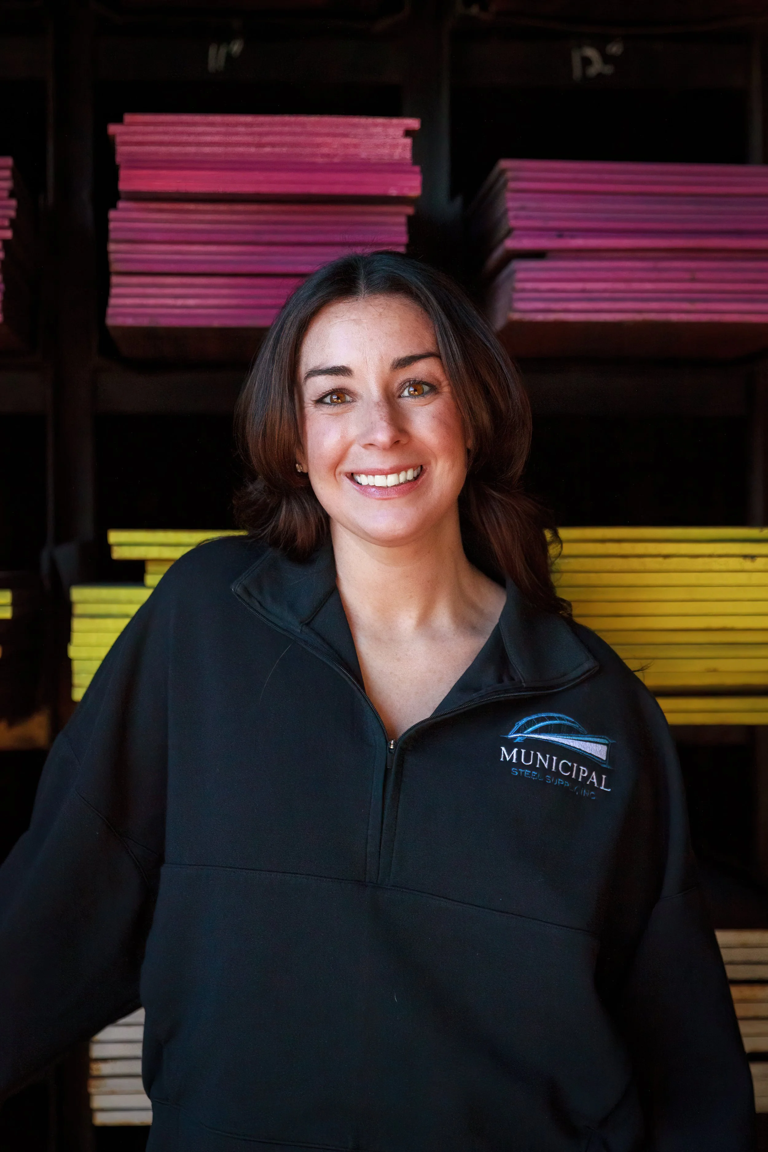 A woman with brown hair and a big smile wearing a black jacket with a municipal logo on it, standing in front of shelves with pink, yellow, and white color samples.