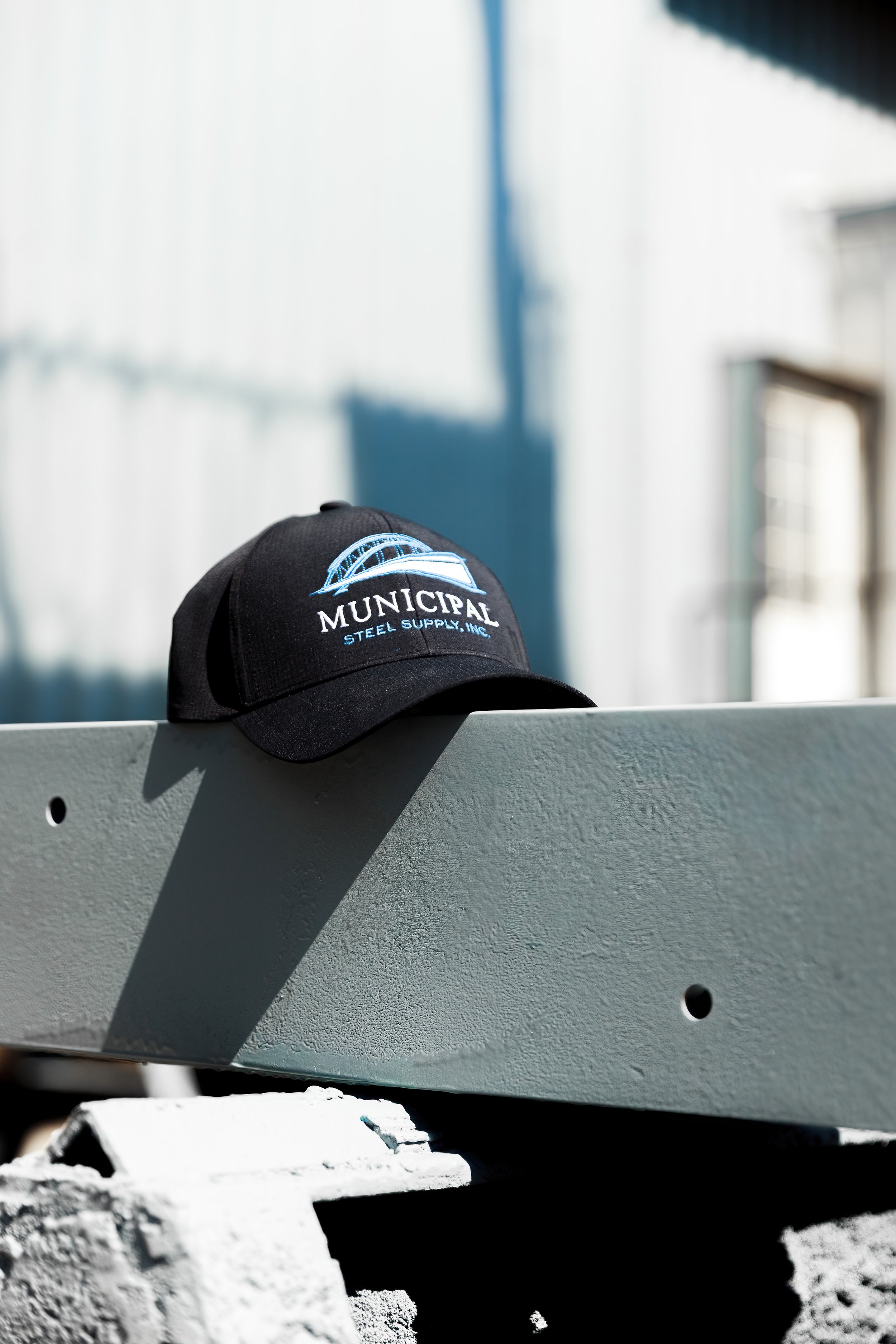 Close-up of a black baseball cap with a embroidered logo of a bridge and the words "Municipal Steel Supply, Inc." resting on a light-colored construction material. The background is blurred, showing a light-colored building and a blue sky.