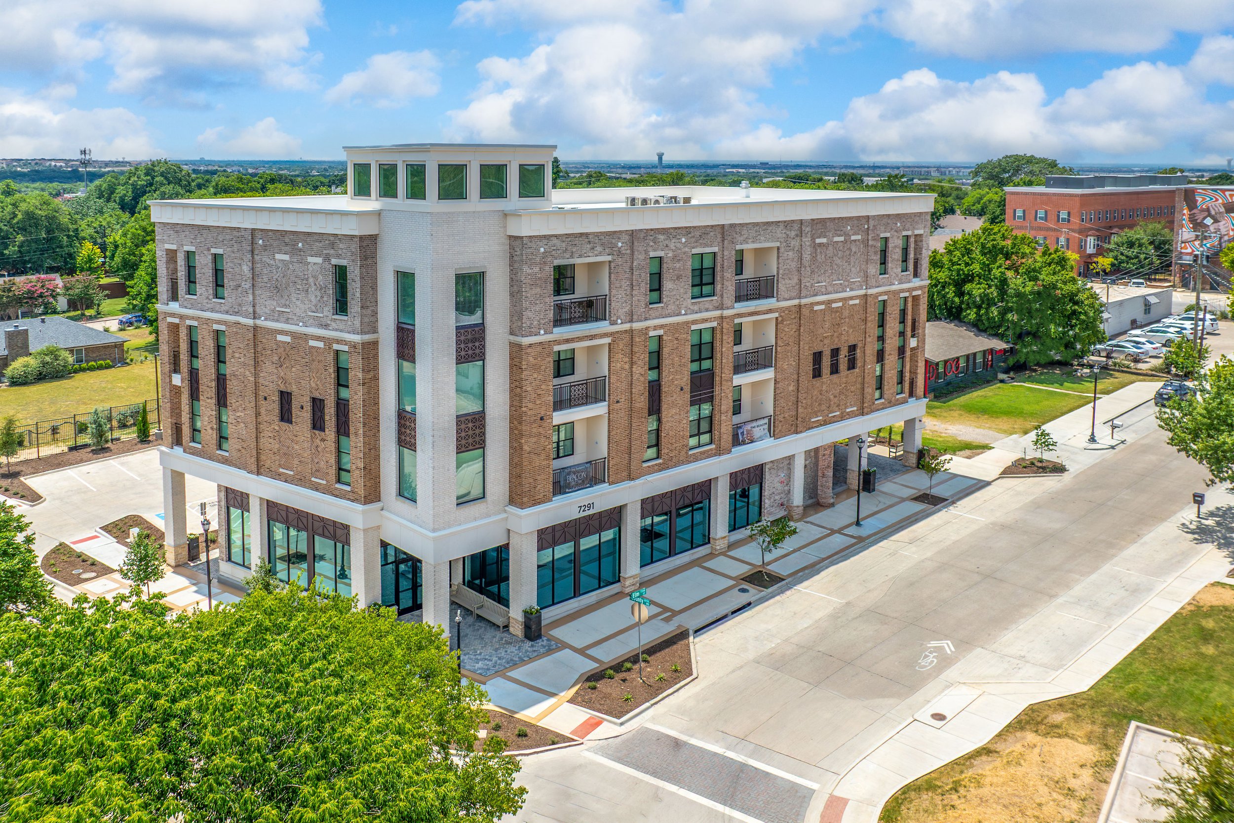 A multi-story residential and commercial building with brick exterior and large windows, situated on a city street corner with trees, sidewalks, and parking.