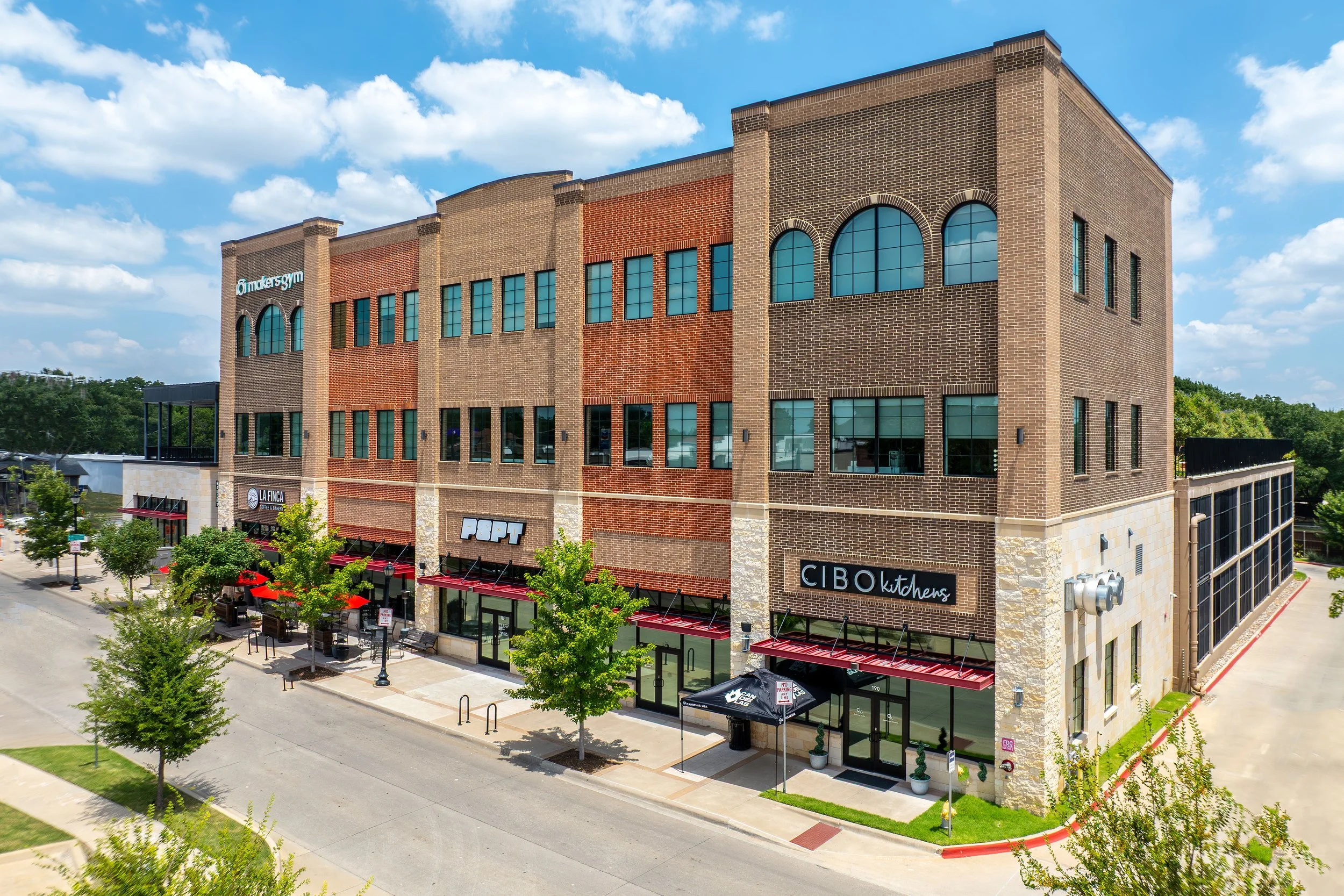 A multi-story commercial building with retail stores on the ground floor, including restaurants and cafes, under a blue sky with scattered clouds. The street in front has trees, street lamps, and outdoor seating areas.