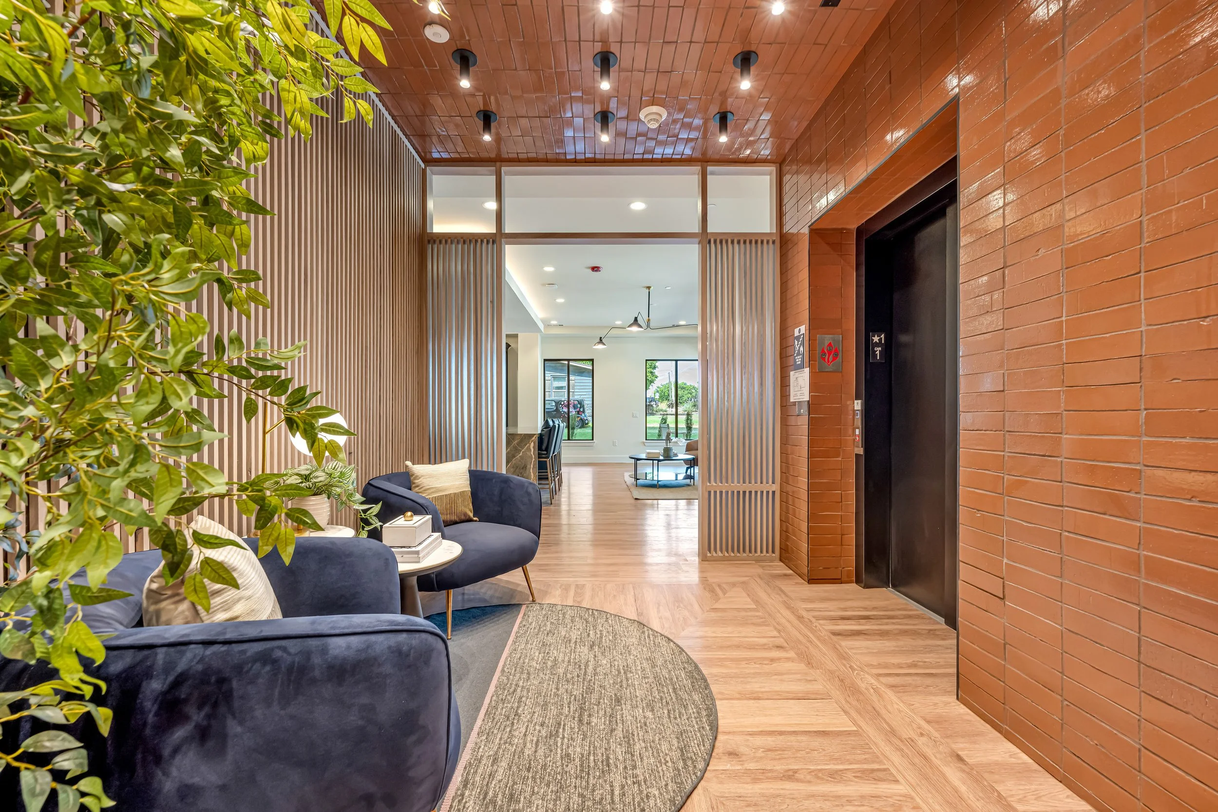 Interior view of a modern building lobby with a seating area featuring navy blue armchairs and pillows, wooden paneled walls, a brick wall next to an elevator door, and visible windows and living space in the background.