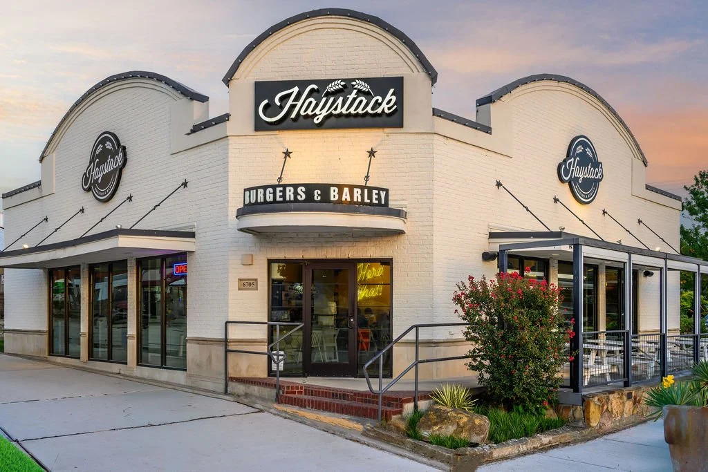 A white brick restaurant named Haystack that serves burgers and barley, with large glass windows, outdoor seating, and decorative plants. The sign above the entrance says 'Haystack' with a smaller sign below that says 'Burgers & Barley'.