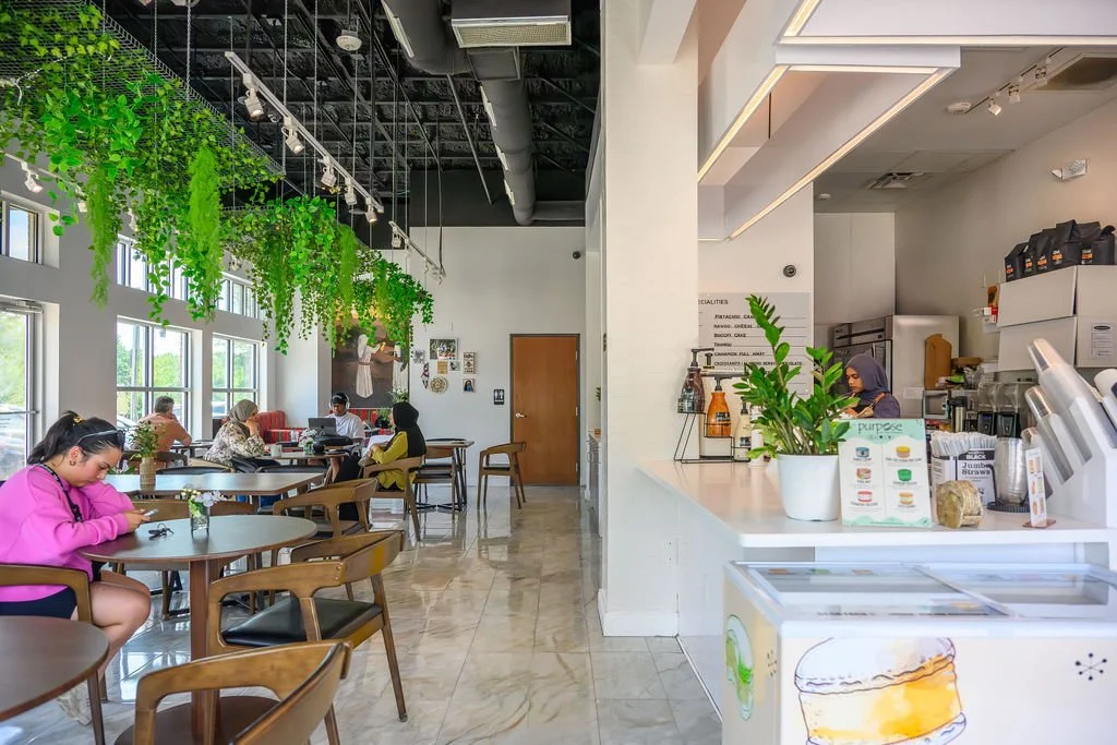 Interior of a modern cafe with hanging green plants, large windows, customers seated at tables, and a counter area with staff preparing drinks.