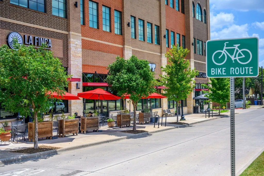 A city sidewalk with outdoor seating under red umbrellas, trees, and a building with a sign for La Fina. A green bike route sign is in the foreground.