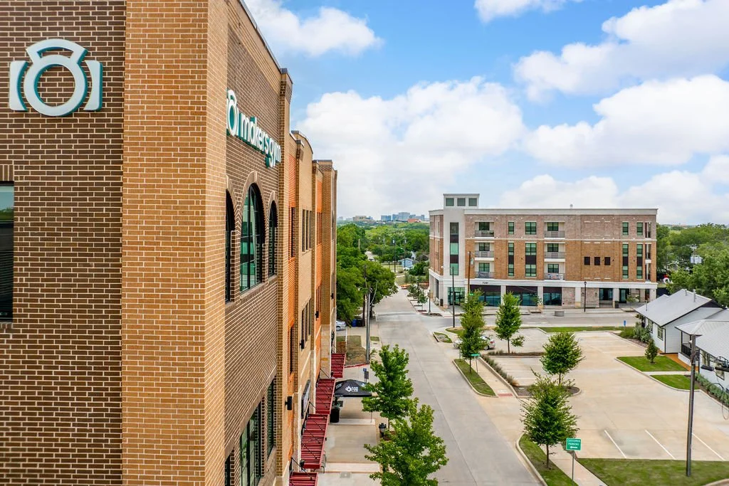 A daytime view of a modern commercial area with brick buildings, parking lot, and green trees, under a partly cloudy sky.
