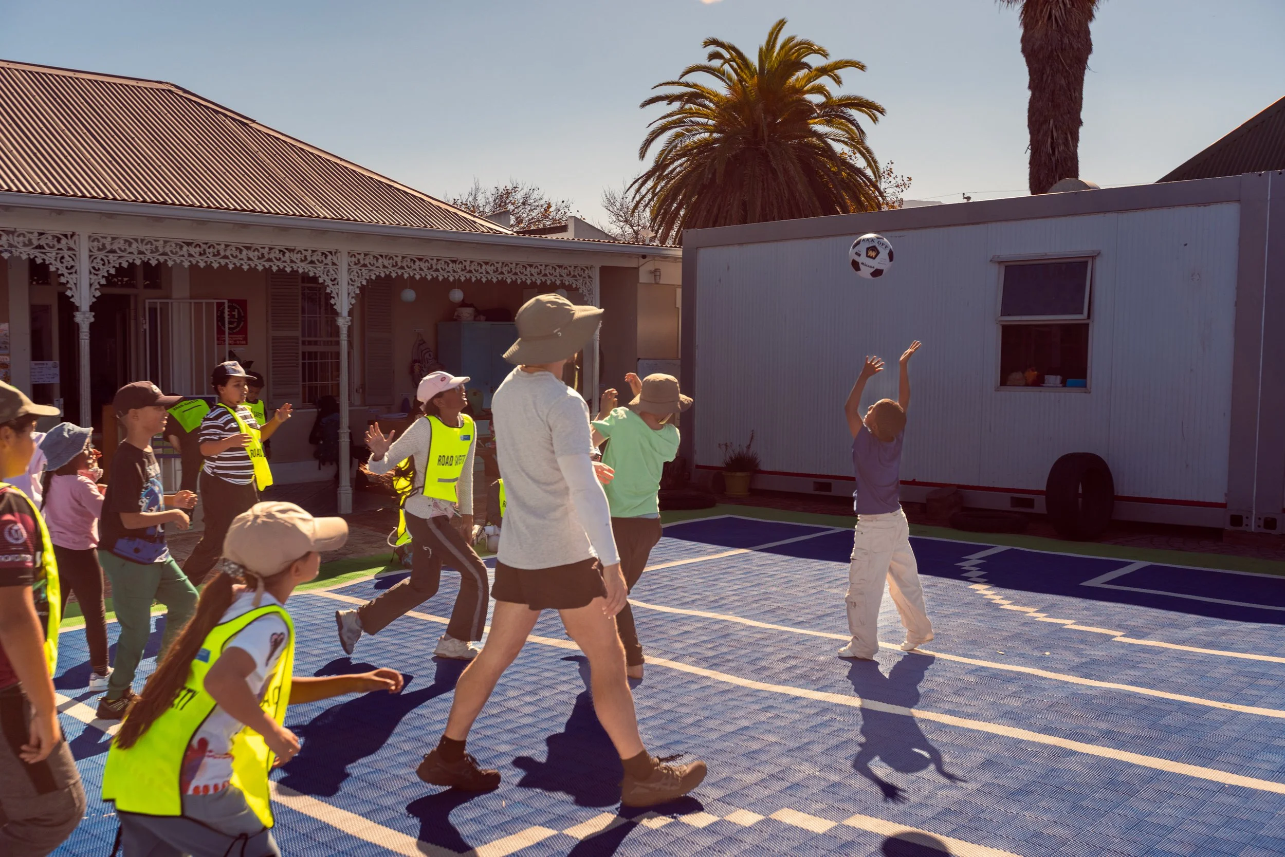 Children and adults playing and watching a game outside, with a soccer ball in the air, in a courtyard with palm trees, a building with decorative white trim, and a trailer in the background.
