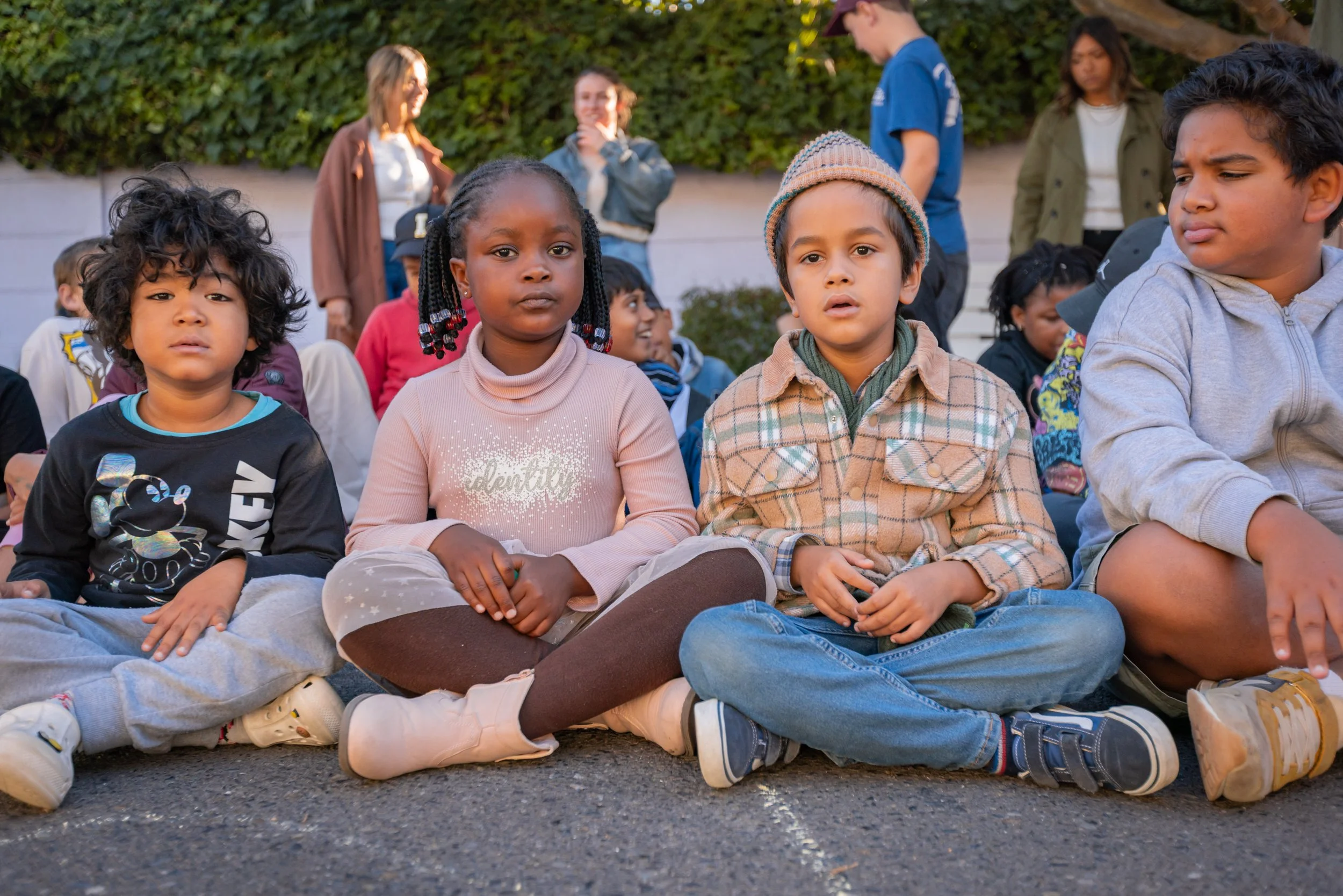 Children sitting on the ground outdoors, some with crossed legs, facing forward, with adults standing and talking in the background. It appears to be an outdoor gathering in a park or similar setting during daytime.