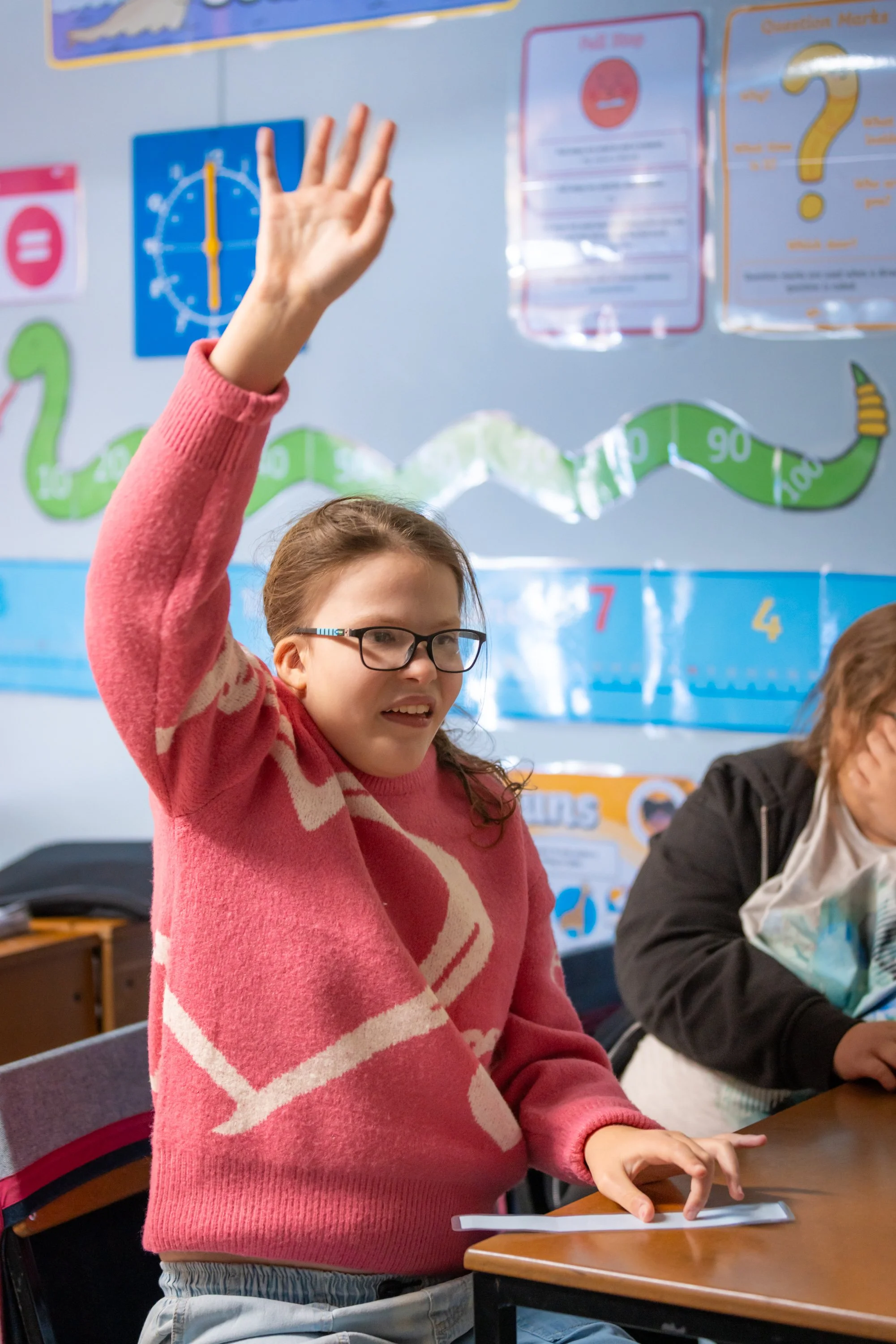 A young girl with glasses and a pink sweater with white patterns raises her hand in a classroom. She is standing next to a desk with a paper on it. Other students and colorful educational posters are visible in the background.