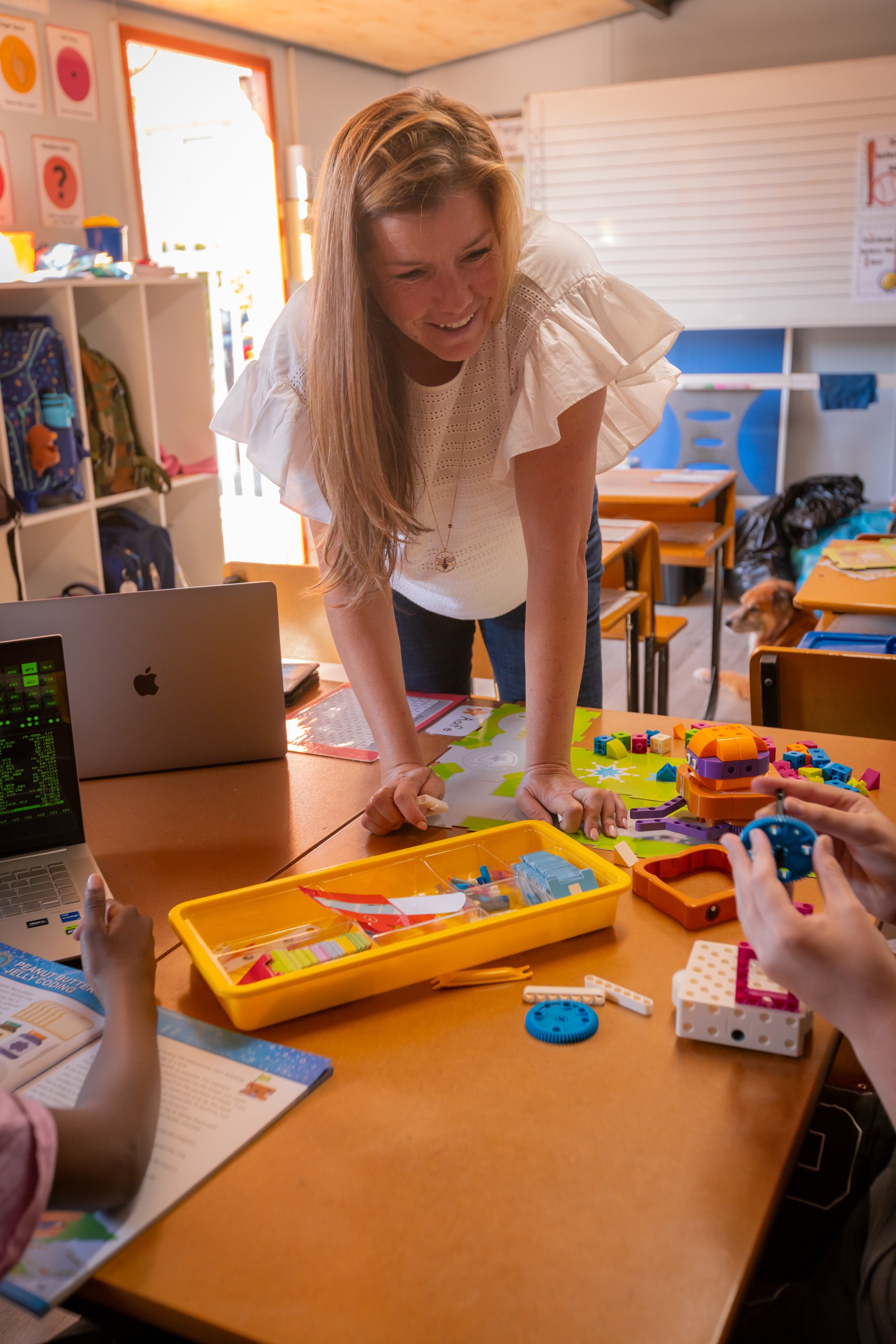 A woman with long blonde hair, wearing a white blouse, leans over a table with her hands on it, smiling, in a colorful classroom with backpacks and educational posters in the background. Several children are working with colorful building blocks and educational toys on the table, with laptops and books nearby.