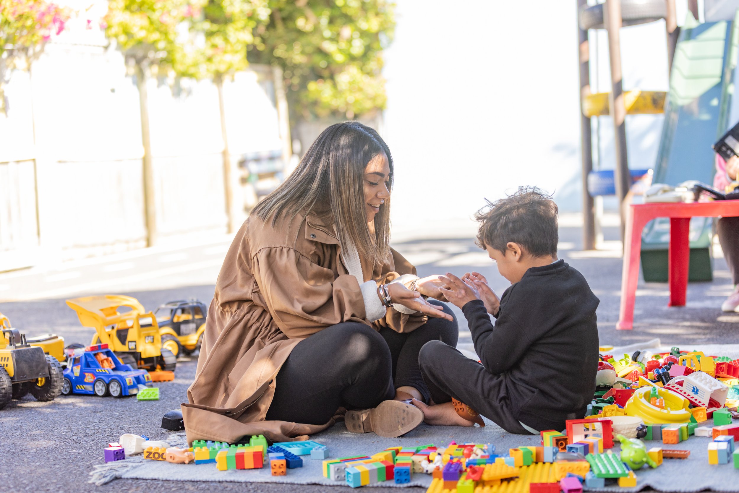 A woman and a young boy playing with colorful toy blocks on the ground outdoors, surrounded by toy vehicles, with trees and a fenced area in the background.