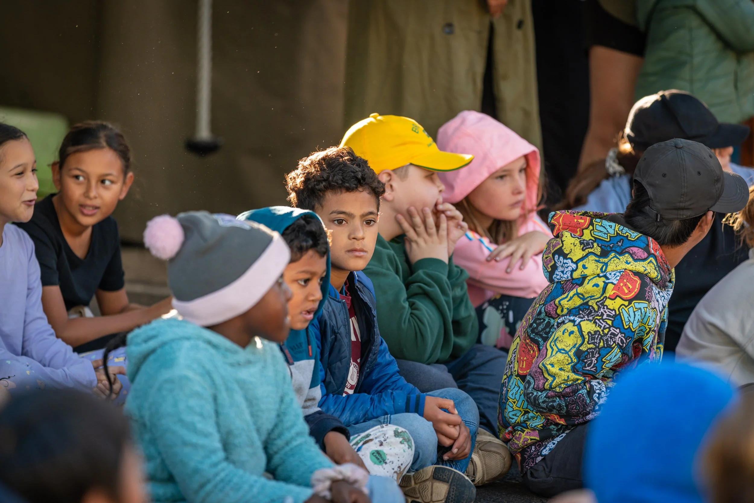 Group of children sitting outdoors, some wearing hoodies and hats, looking attentively in a specific direction.
