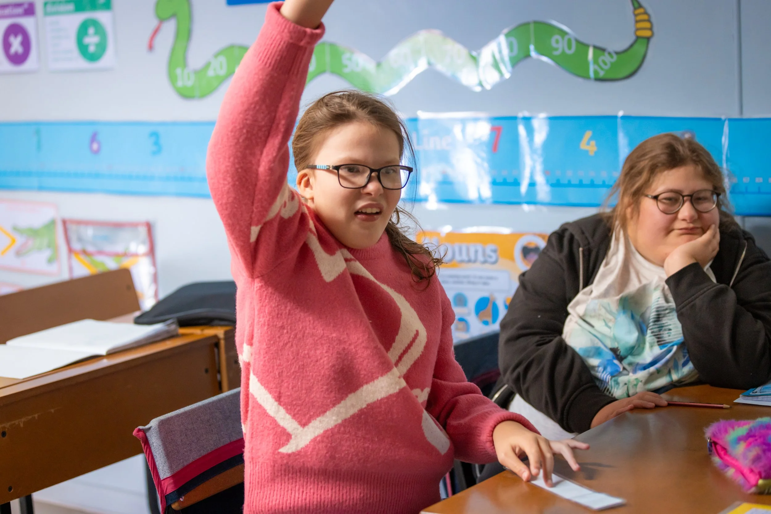 A girl in a pink and white striped sweater raising her hand in a classroom, with another girl sitting next to her resting her chin on her hand, and educational posters and a class schedule on the wall behind them.