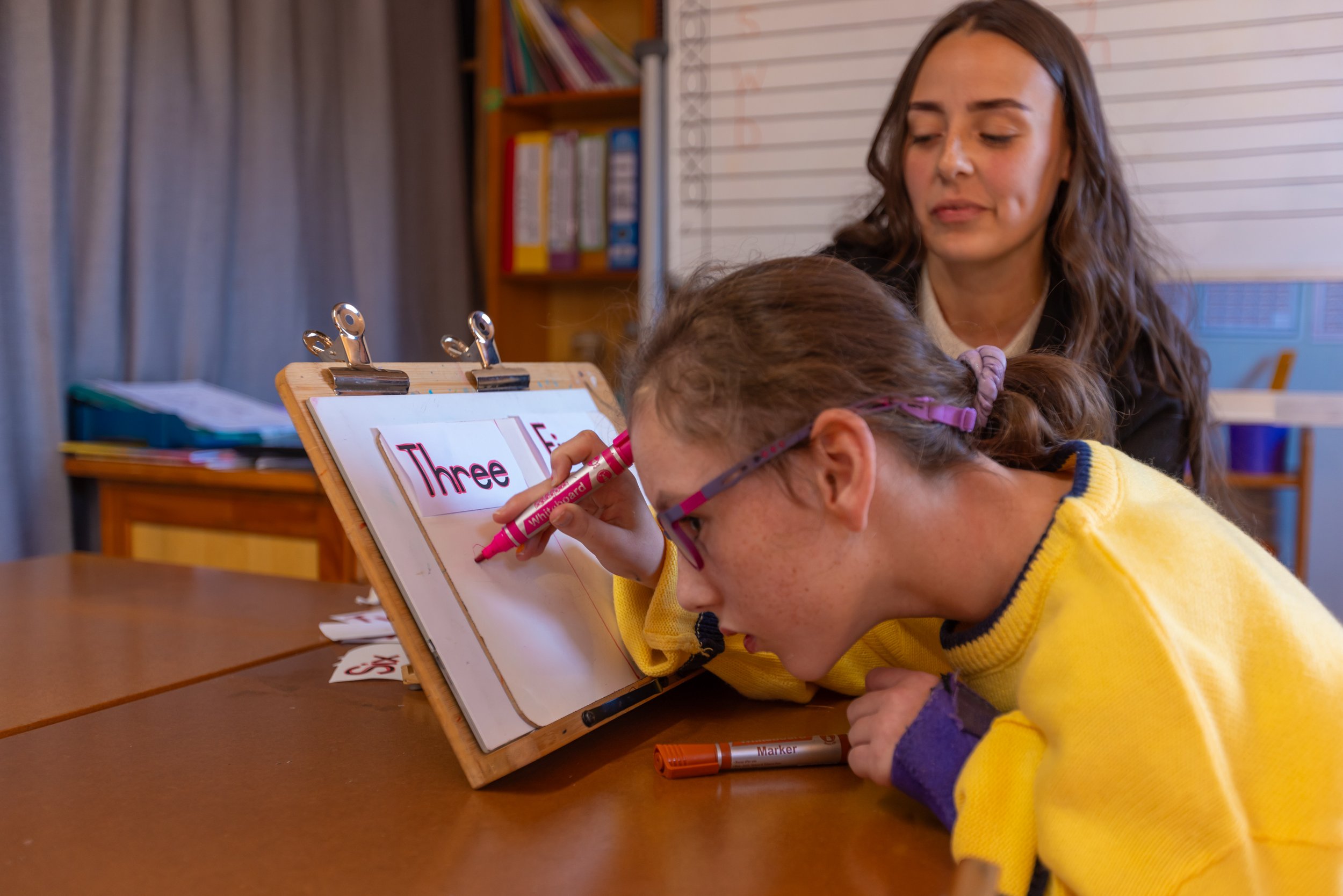 A teacher with long brown hair and a student with blonde hair and glasses, both at a table in a classroom. The student is writing the word 'Three' on a whiteboard with a pink marker, while the teacher looks on. There are bookshelves and educational m