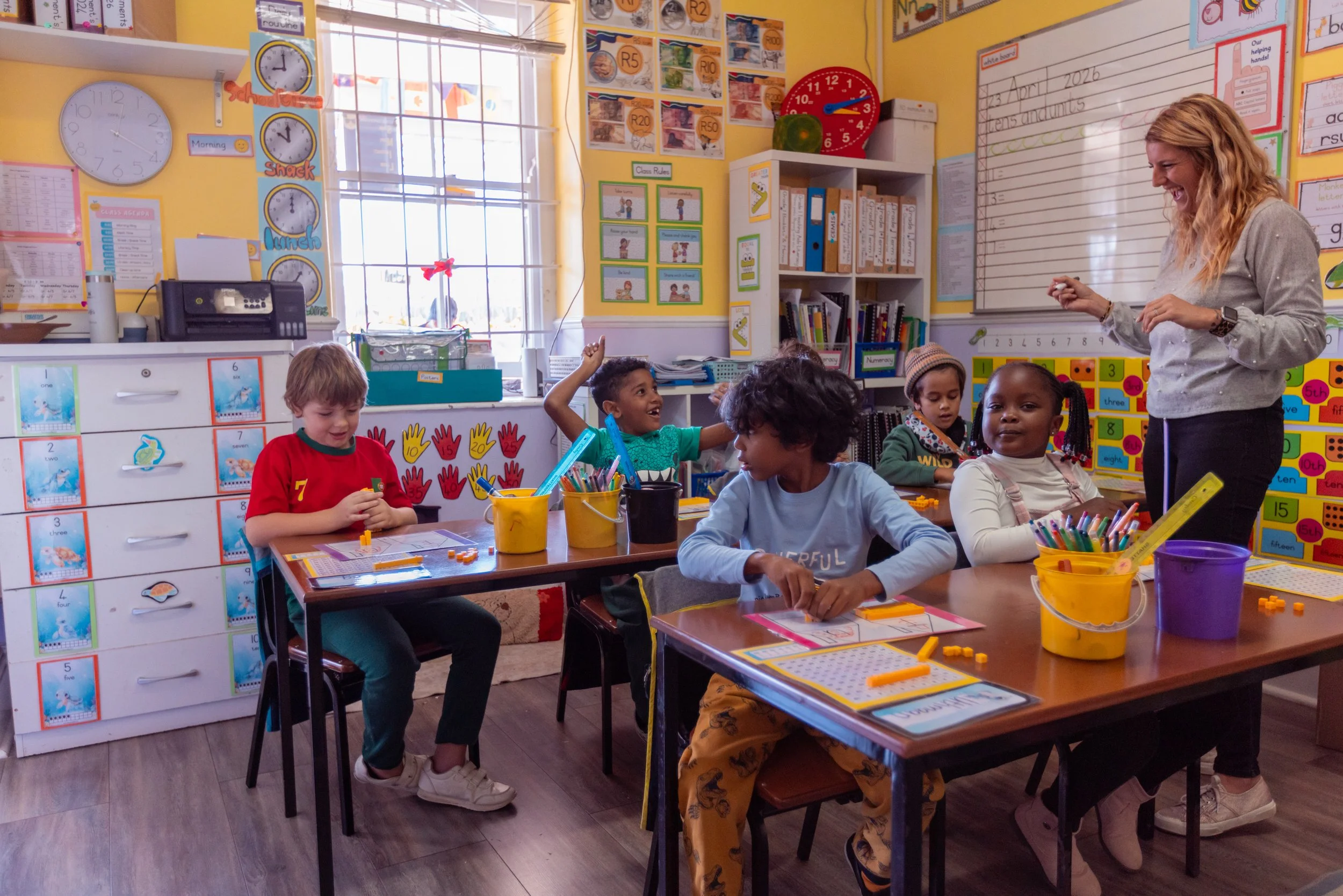 A classroom with young children sitting at tables, engaged in activities. A teacher stands nearby, smiling and interacting with students. The room is colorful with educational posters, clocks, and shelves filled with books and supplies.
