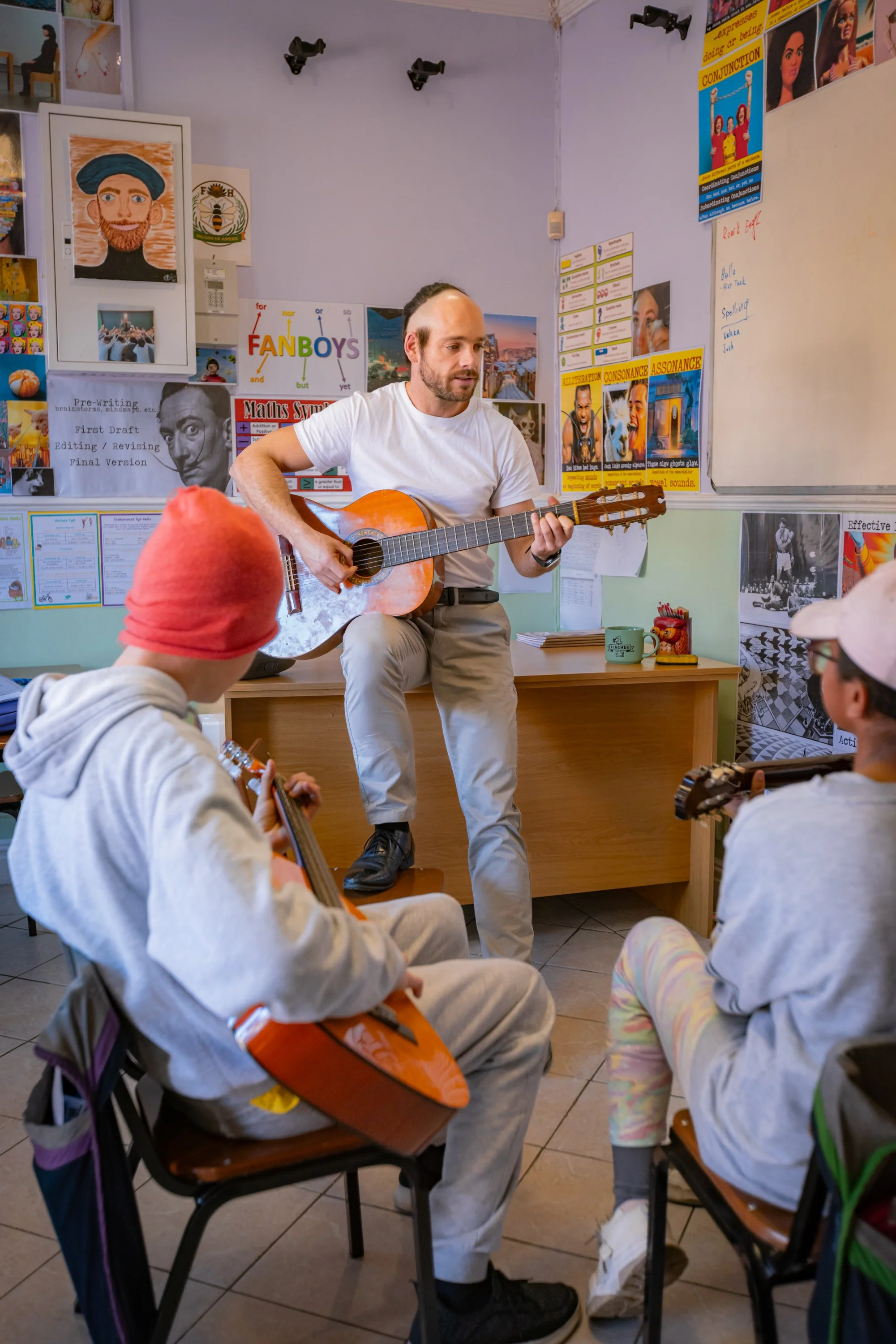 A man with a guitar performing for children seated in a classroom, with posters, pictures, and colorful decorations on the walls.