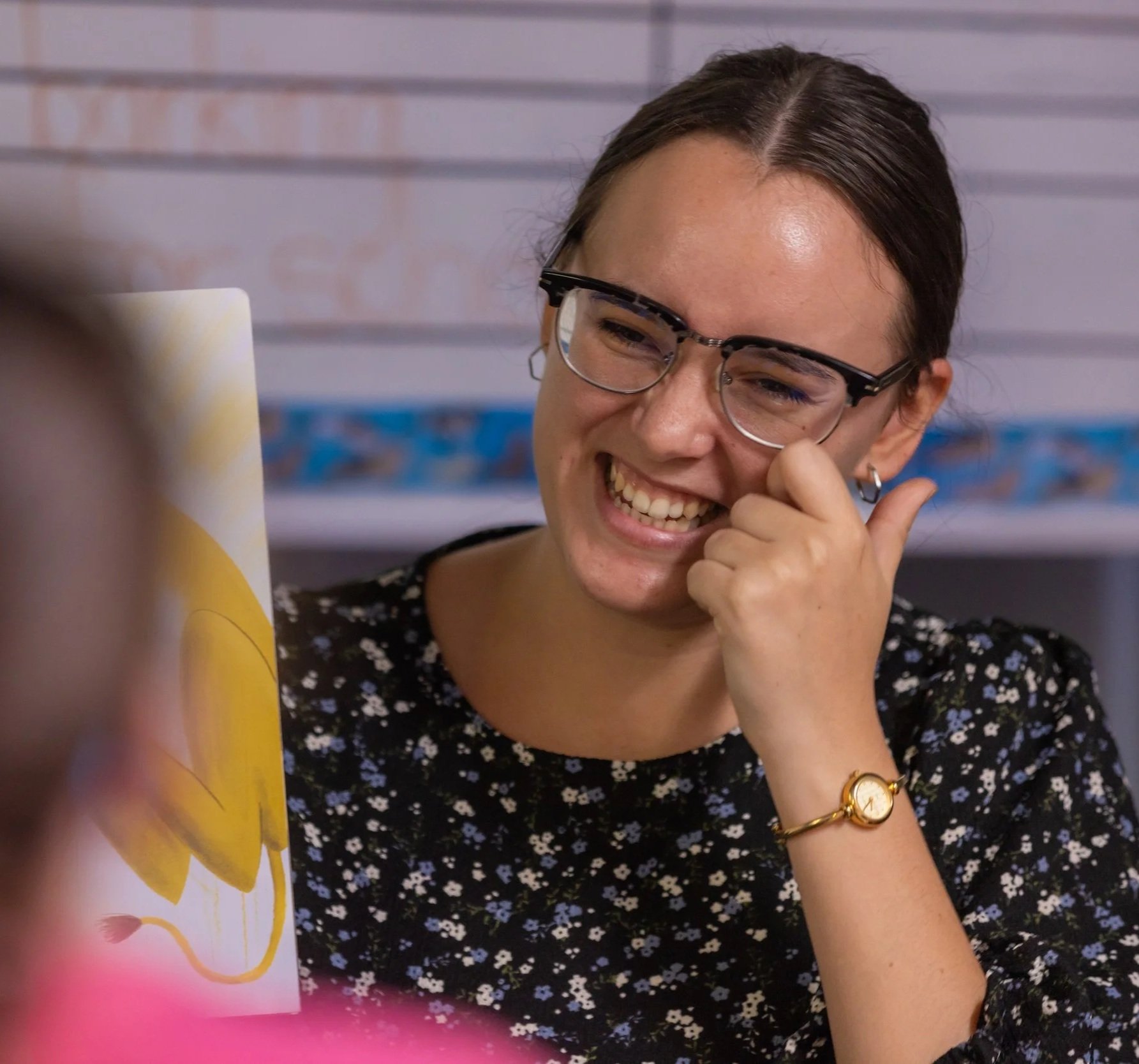A woman with short dark hair and glasses is smiling and touching her face, sitting in front of a laptop with a patterned background.
