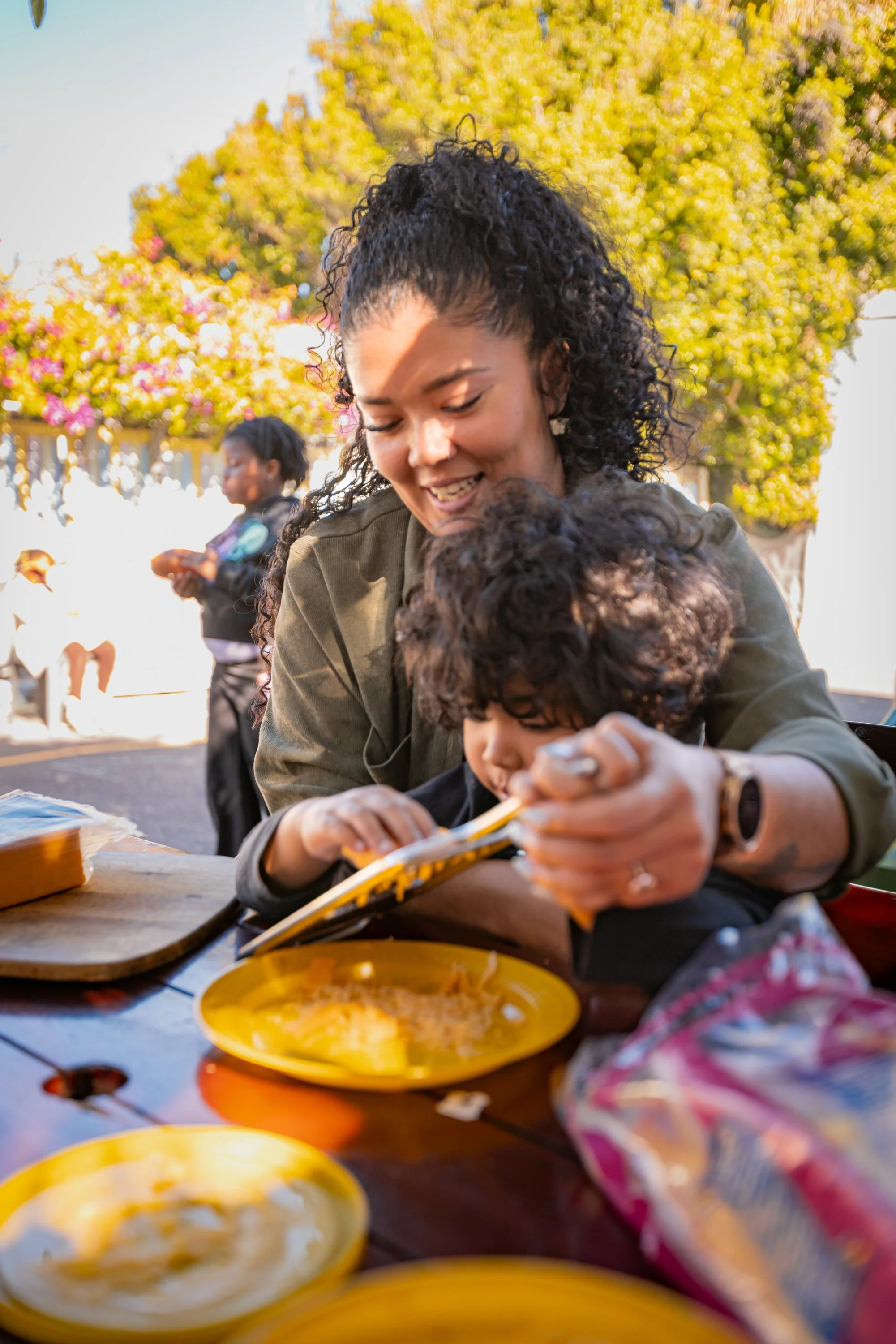 A woman with curly black hair smiling and hugging a child with curly brown hair at an outdoor gathering. There are yellow plates with food on a wooden table and other children in the background with yellow and pink blooming trees.