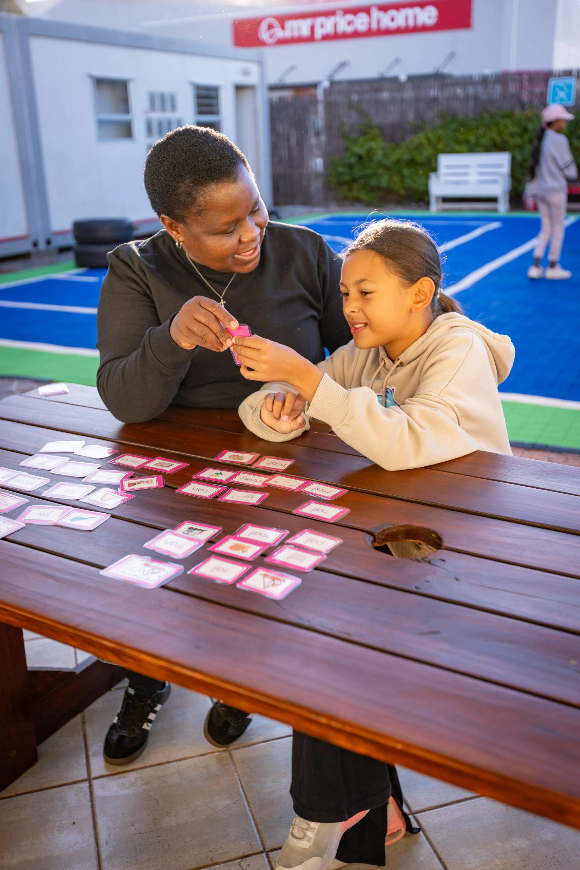 A woman and a girl are sitting at a wooden table outdoors, playing a vocabulary card game. The woman, with short hair and wearing a black shirt, is handing a card to the girl. The girl has long brown hair and is wearing a beige hoodie. There are multiple cards on the table with words and pictures. In the background, there is a blue tennis court, some benches, a fence, and a woman standing near the fence. A sign for Price Home is visible above them.
