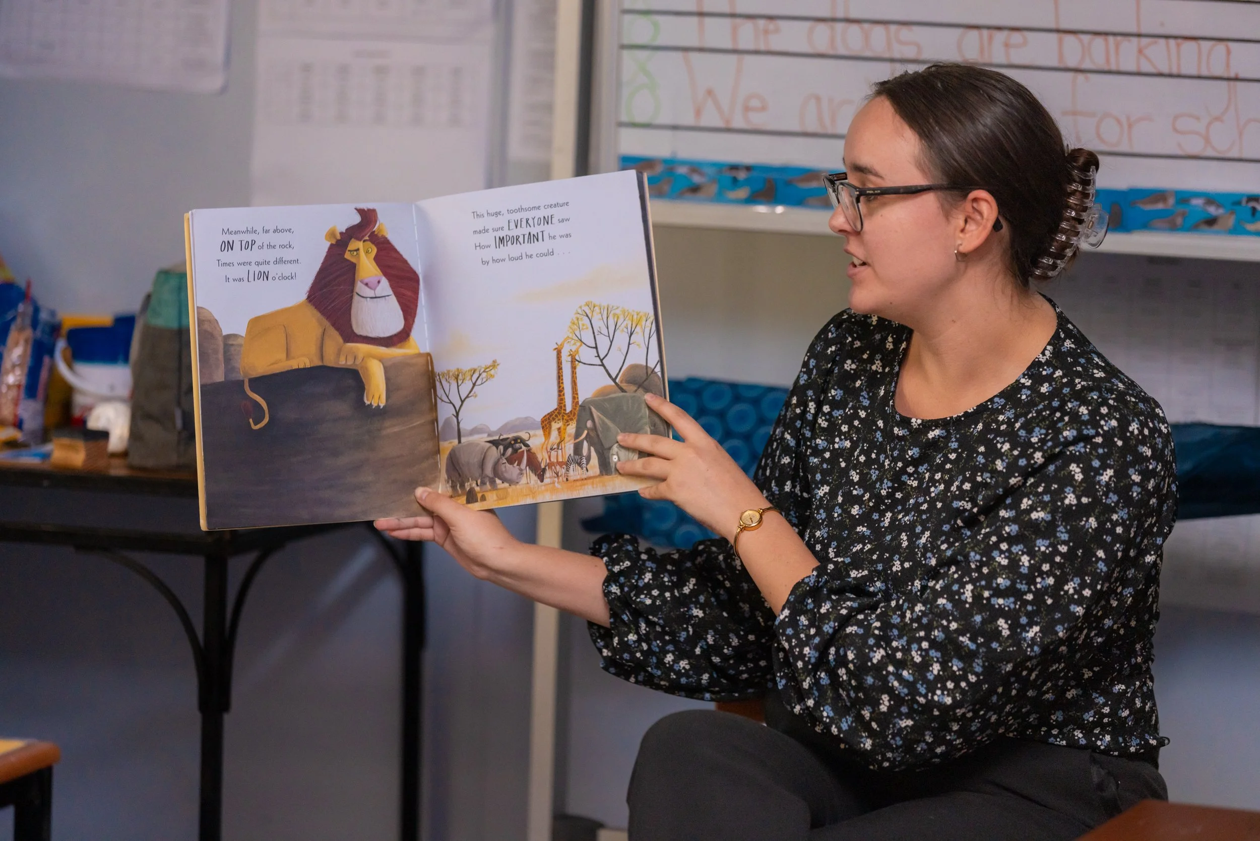 A woman with glasses and a watch, wearing a black floral blouse, is sitting and reading a children's picture book with illustrated animals, including a lion, giraffe, and zebra, in a classroom setting.