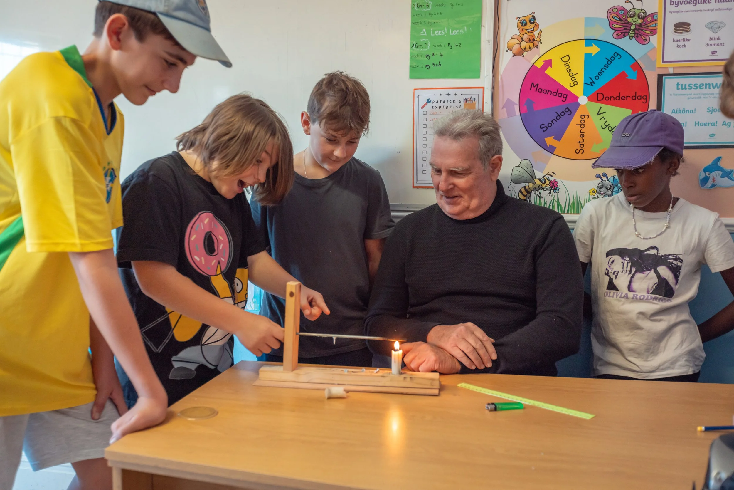 Children and an adult gathered around a table with a candle and a wooden stand, engaging in a science experiment or demonstration in a classroom with colorful educational posters in the background.