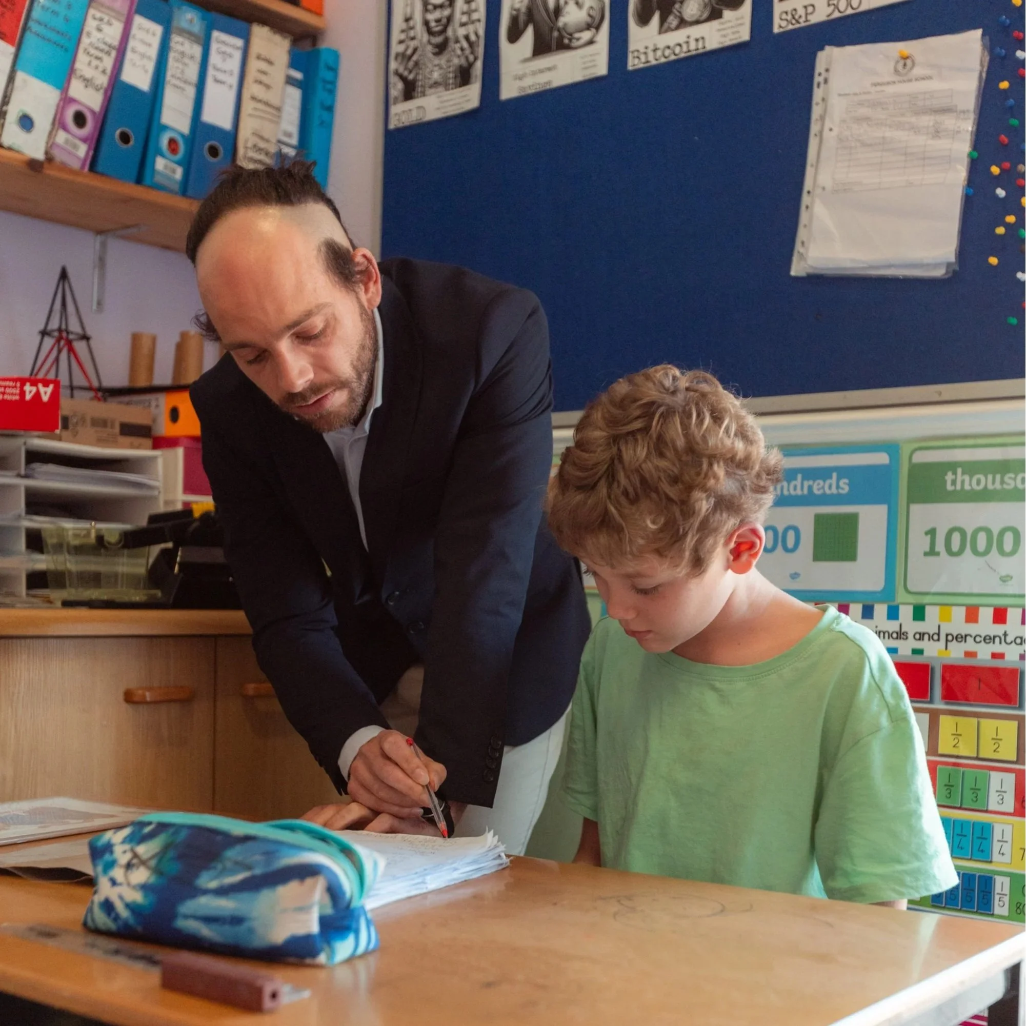 A man in a dark suit looking down at papers on a classroom desk next to a child with curly hair wearing a green shirt. The classroom has educational posters, binders, and a bulletin board on the walls.