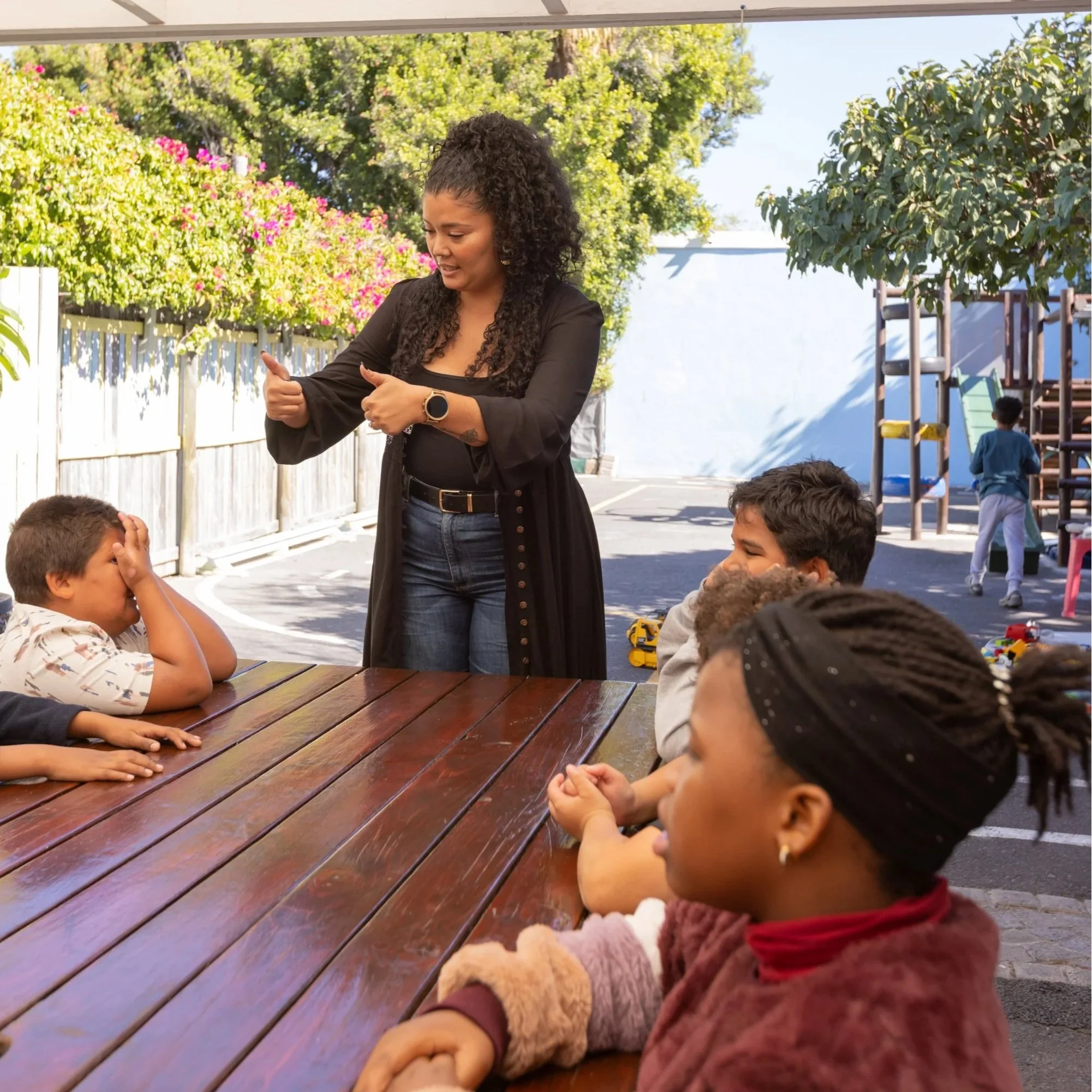 A woman with curly hair wearing a black jacket and jeans is standing outdoors, engaging with children around a wooden picnic table. She appears to be talking or explaining something to the children. In the background, there are colorful flowers, green trees, and playground equipment on a bright, sunny day.