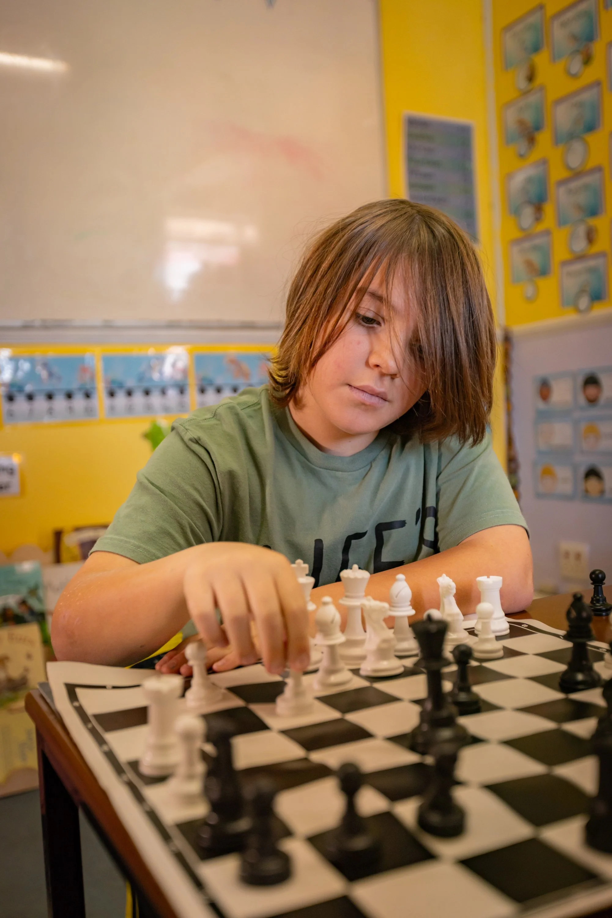 A young boy with shoulder-length brown hair playing chess in a classroom with yellow walls and educational posters.