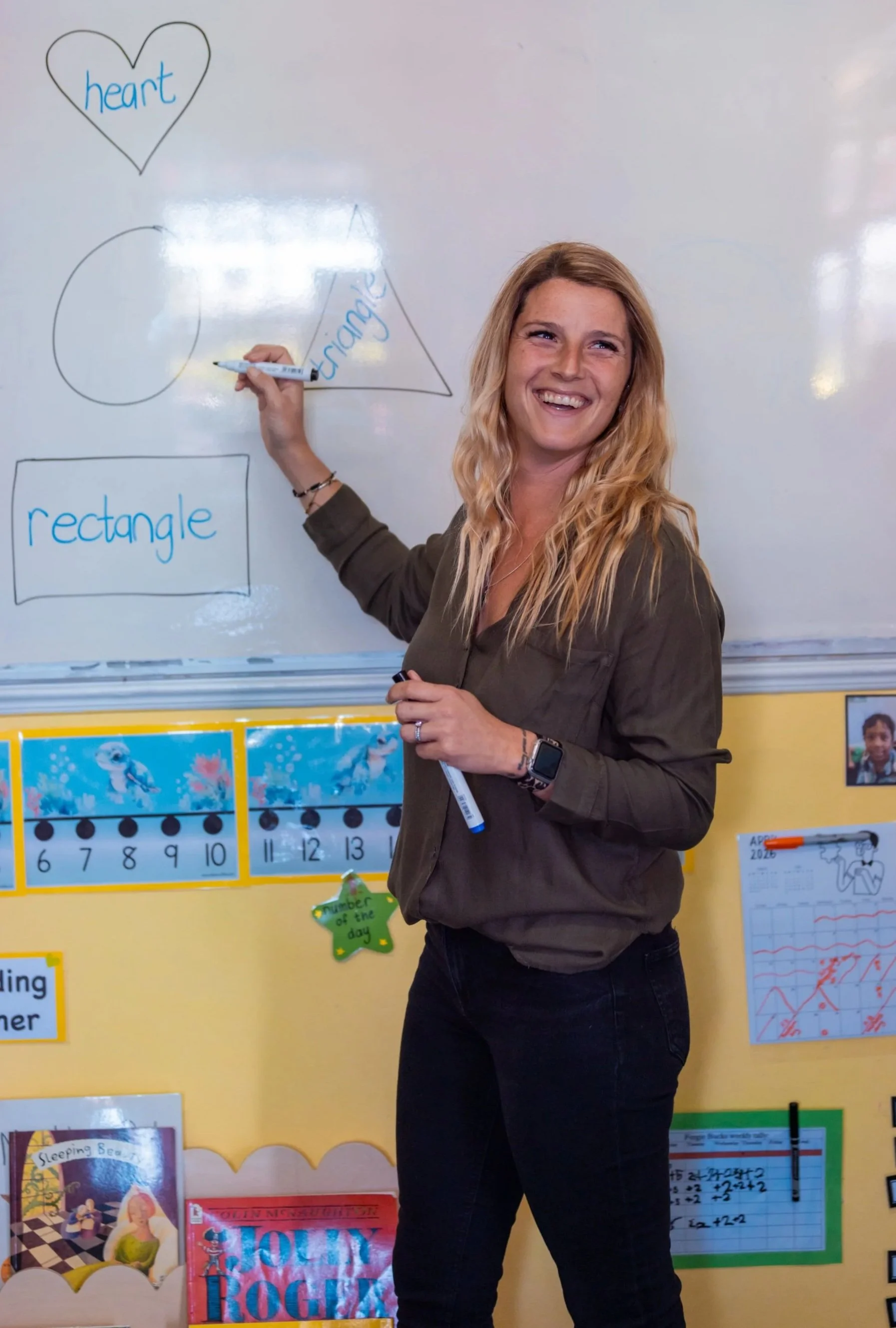 A woman with long wavy blonde hair smiling and holding a dry erase marker while standing in front of a whiteboard with drawings of a heart, a triangle, a rectangle, and a circle, each labeled with the respective shape.