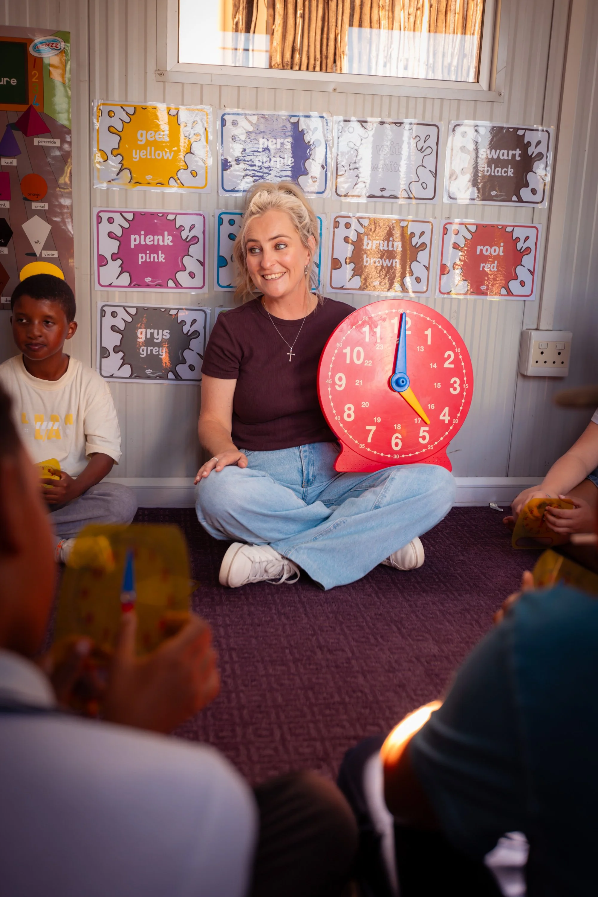 A classroom scene with children sitting on the floor, some holding yellow cards with colorful illustrations. A smiling woman sits cross-legged in front of them, holding a large red clock showing approximately 12:01. Behind her are educational posters