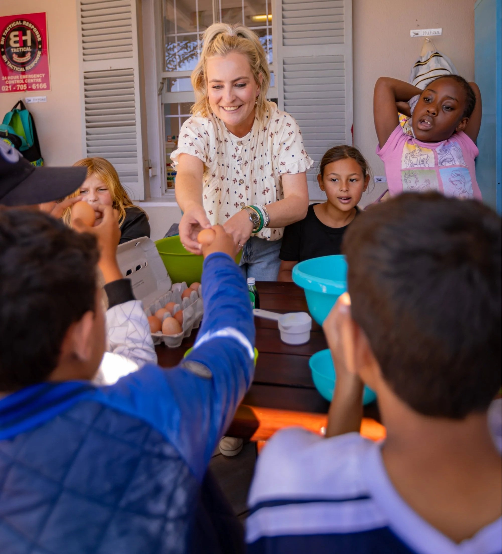 A smiling blonde woman handing an egg to a child at a table, with other children and colorful bowls nearby, in a brightly lit room with shutters and a window in the background.
