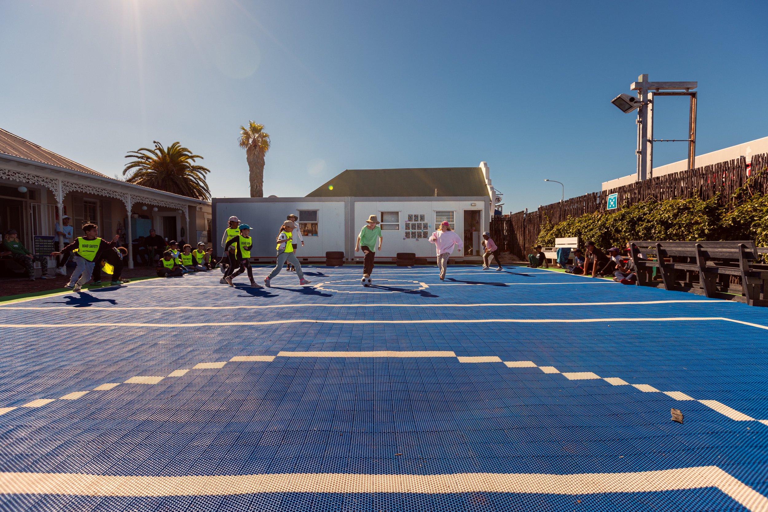 Children playing soccer on a blue outdoor court with onlookers sitting along the side, surrounded by buildings and palm trees, under a clear sunny sky.